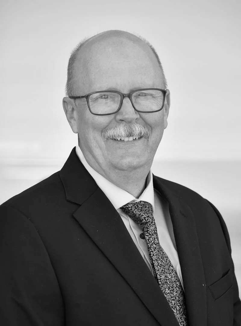 Black and white portrait of a smiling middle-aged man wearing glasses, a suit, tie, and collared shirt.