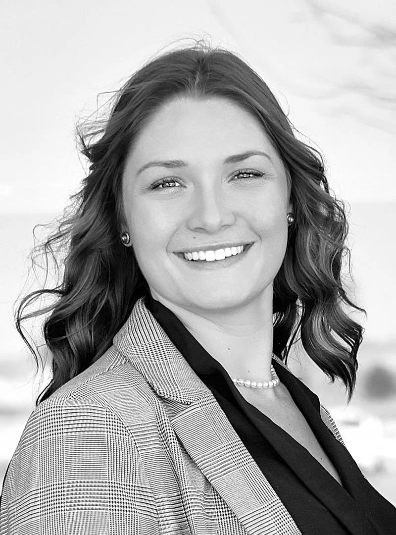 A woman smiling with wavy hair, wearing a blazer, pearl earrings, a necklace, and a black top, outdoors in black and white.
