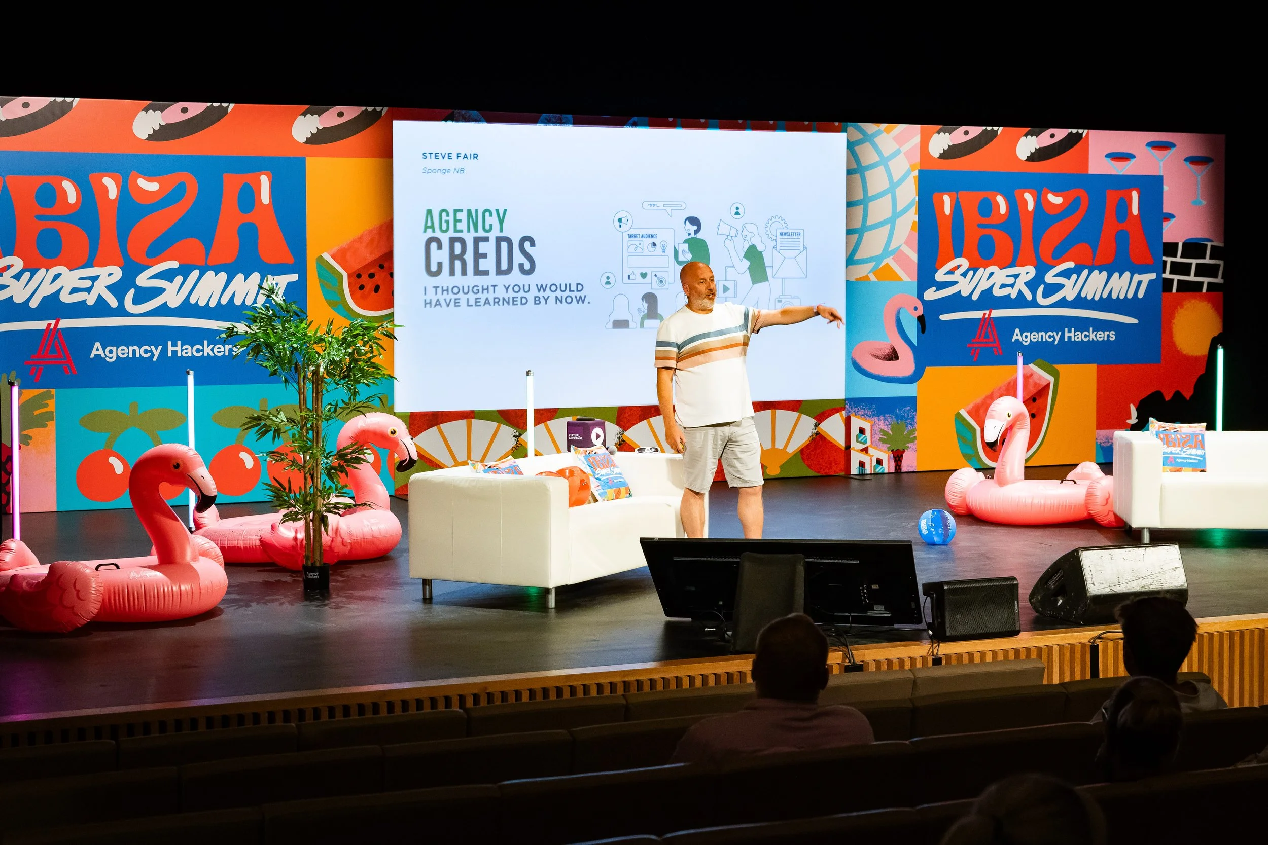 Steve Fair on stage presenting at Agency Hackers Super Summit, with tropical-themed decorations including inflatable flamingos, a palm tree, colourful banners, and a large screen displaying his presentation.