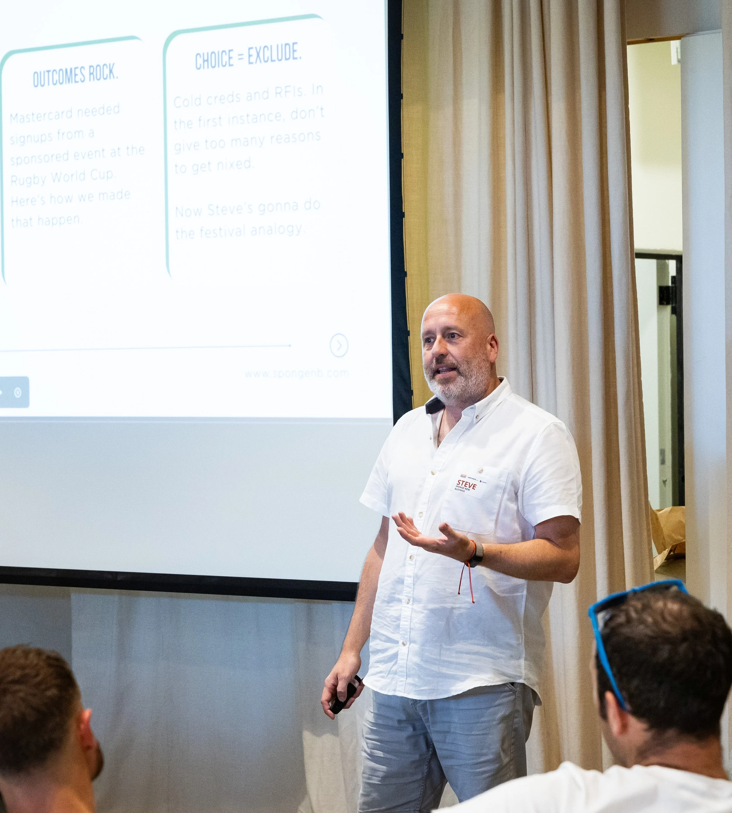 Man with a beard and bald head giving a presentation in front of a screen, with audience members in the foreground, in a room with beige curtains.