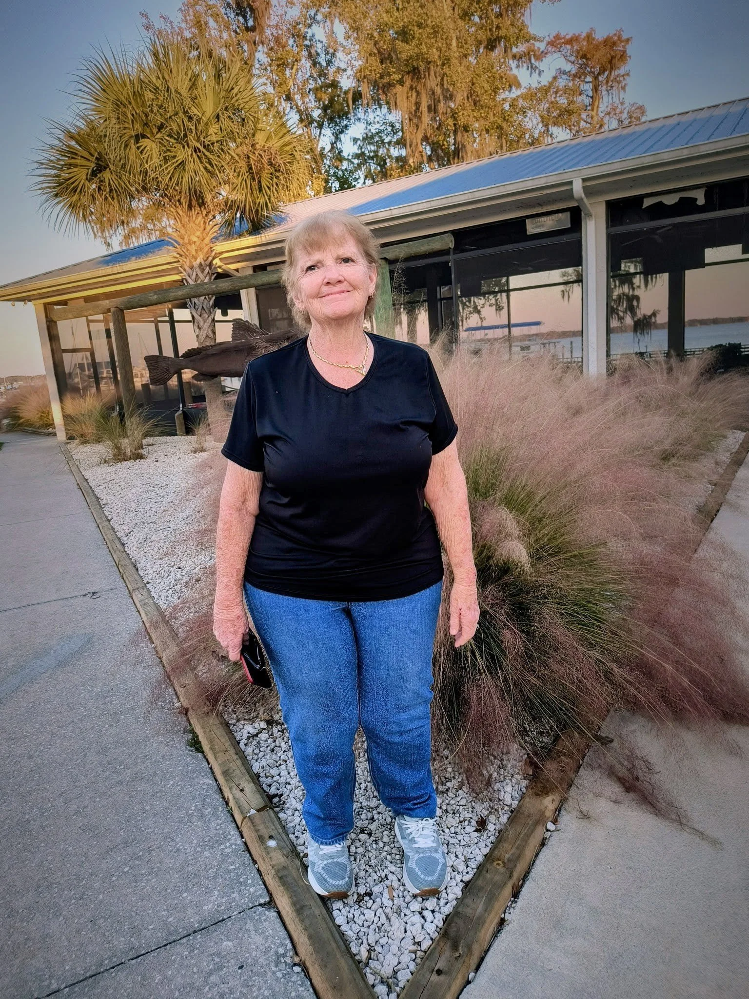 An elderly woman standing outdoors near a building with large windows, palm trees, and ornamental grasses, during sunset.