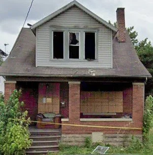 A damaged two-story house with a broken window on the upper floor and boarded-up windows and doors on the ground floor.