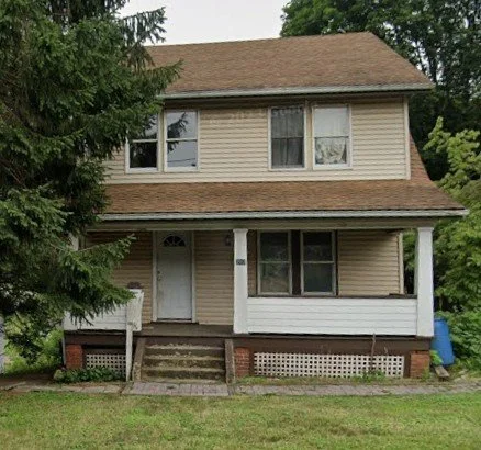 Front view of a two-story house with beige siding, white trim, a front porch, and a gabled roof, surrounded by greenery.
