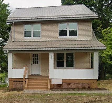 A two-story beige house with a metal roof, front porch, and three steps leading up to the front door, surrounded by a grassy yard and trees in the background.
