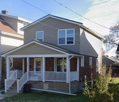 Two-story house with a front porch, white railing, and beige siding, situated in a residential neighborhood.
