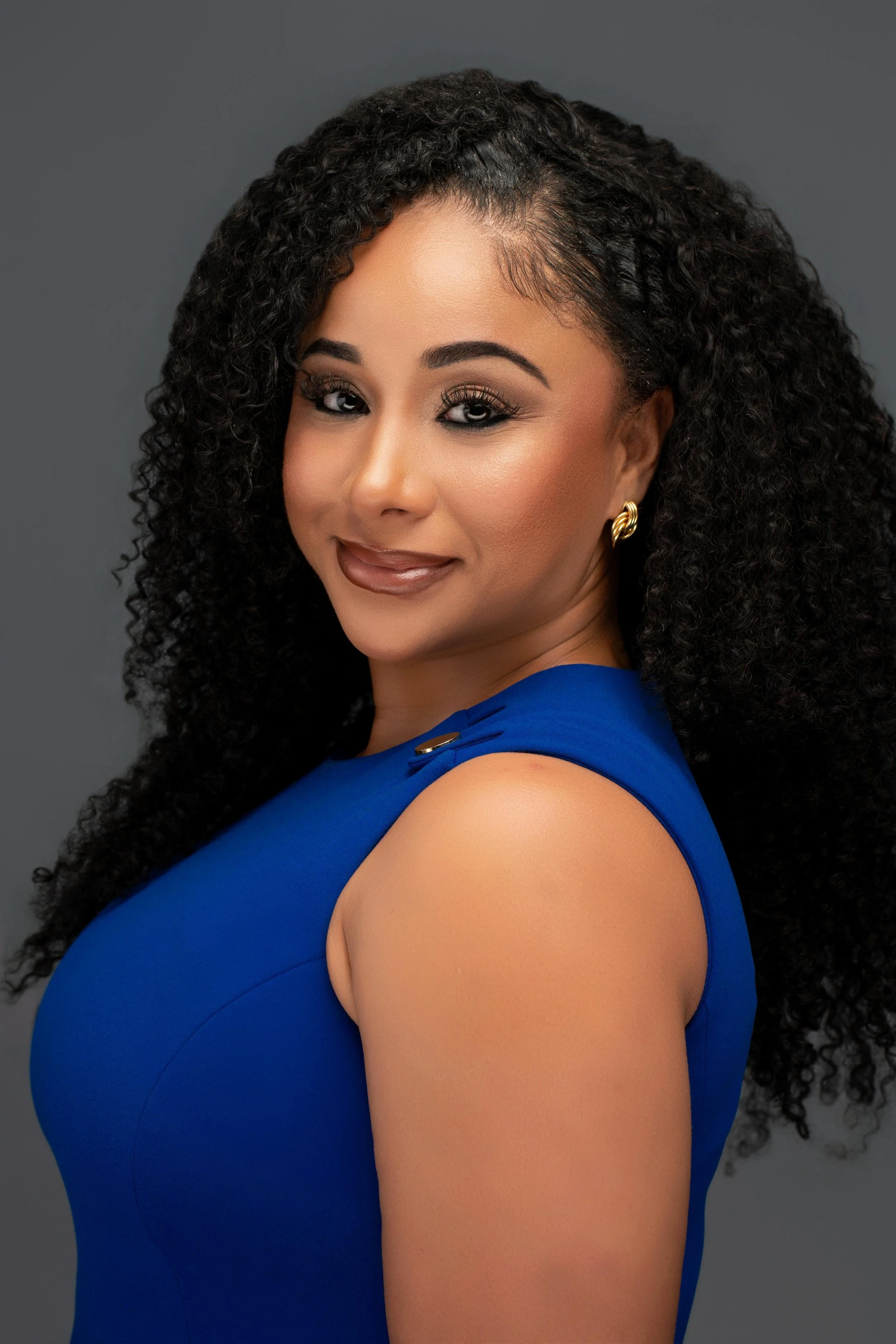 A woman with dark, curly hair wearing a blue sleeveless top and gold earrings against a gray background.