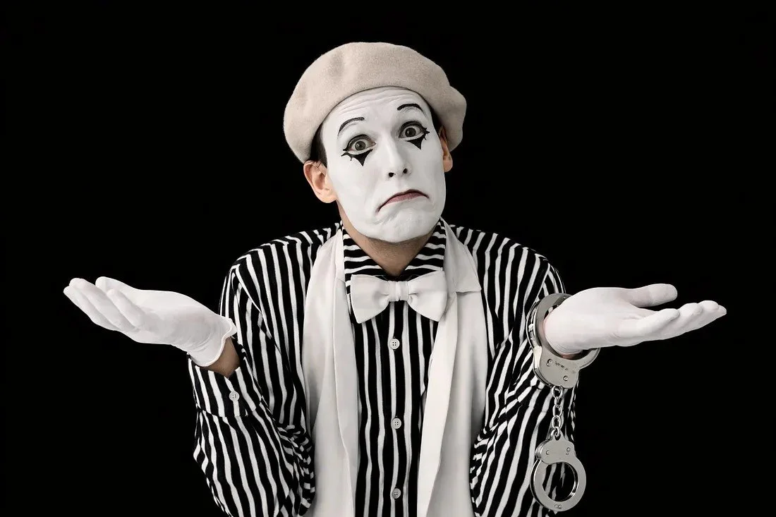 A mime artist dressed in black and white striped shirt, white gloves, and a beige beret, with white face paint and black makeup around the eyes, looks confused with hands raised in a shrug gesture against a black background.