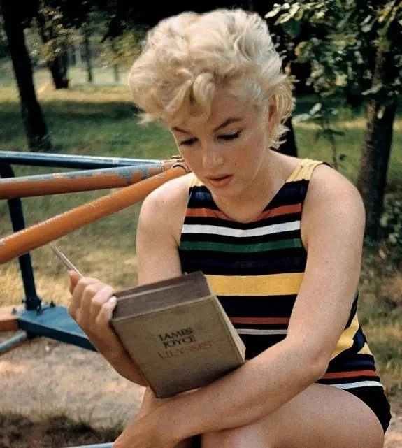 A woman with short, curly blonde hair wearing a sleeveless striped top sits outside in a park, reading a book.