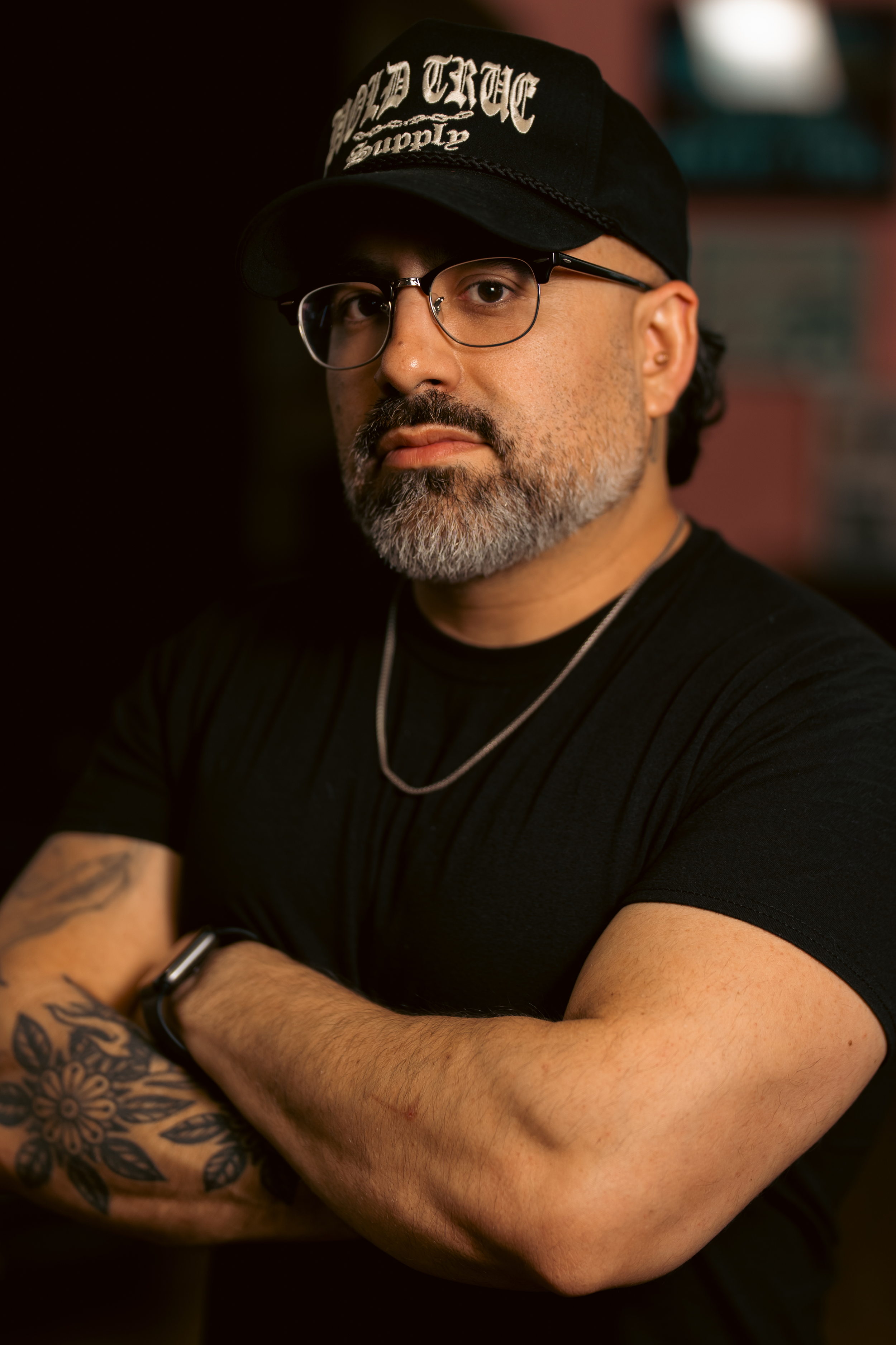 A man with glasses, a beard, and tribal tattoos on his arm wearing a black T-shirt, a chain necklace, and a black cap with white writing, posing with arms crossed in a dimly lit environment.
