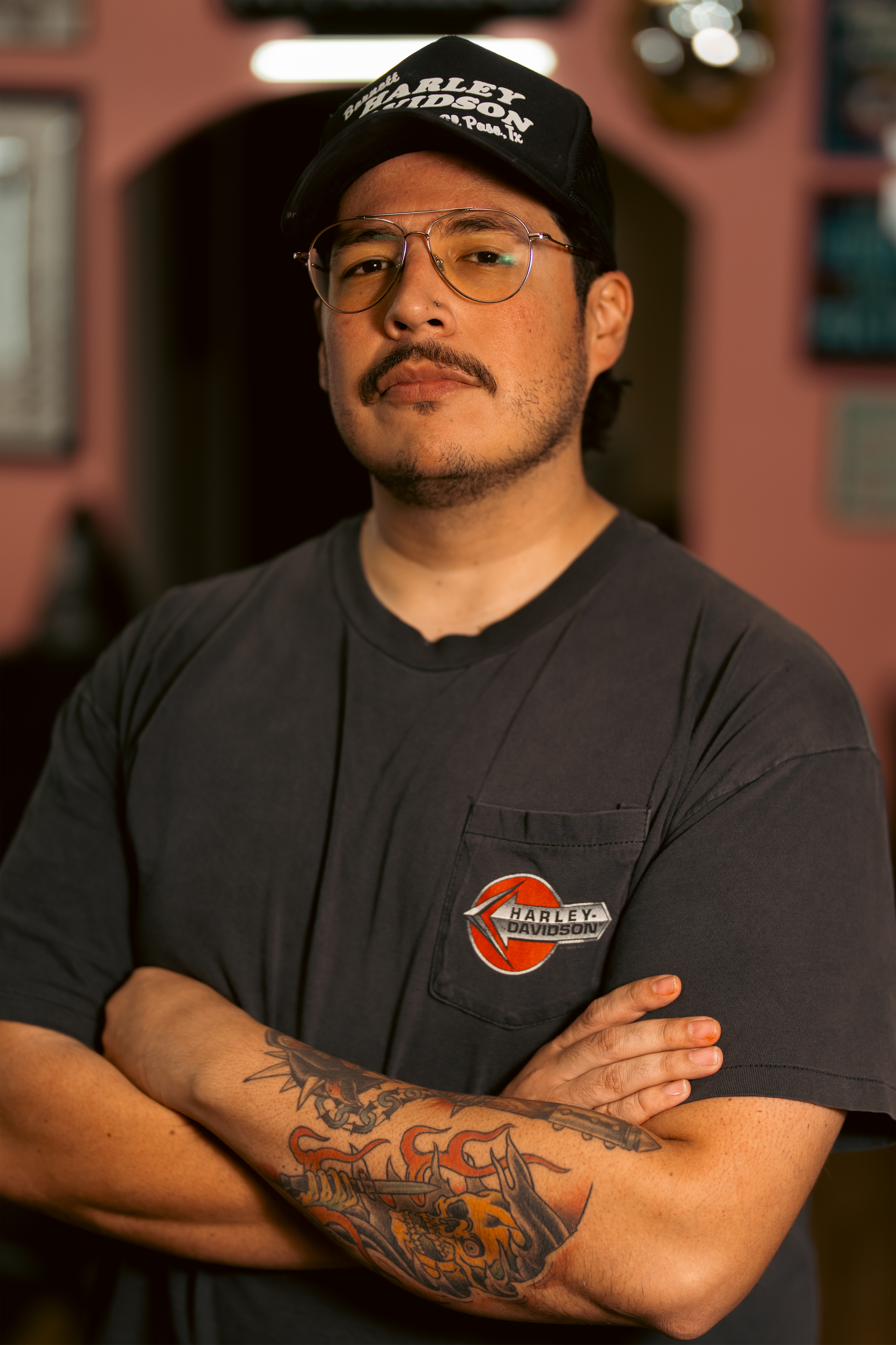 A man with glasses, tattoos on his arm, and a mustache, wearing a black Harley Davidson t-shirt and a black cap, standing with his arms crossed in a room with pink walls and framed pictures.