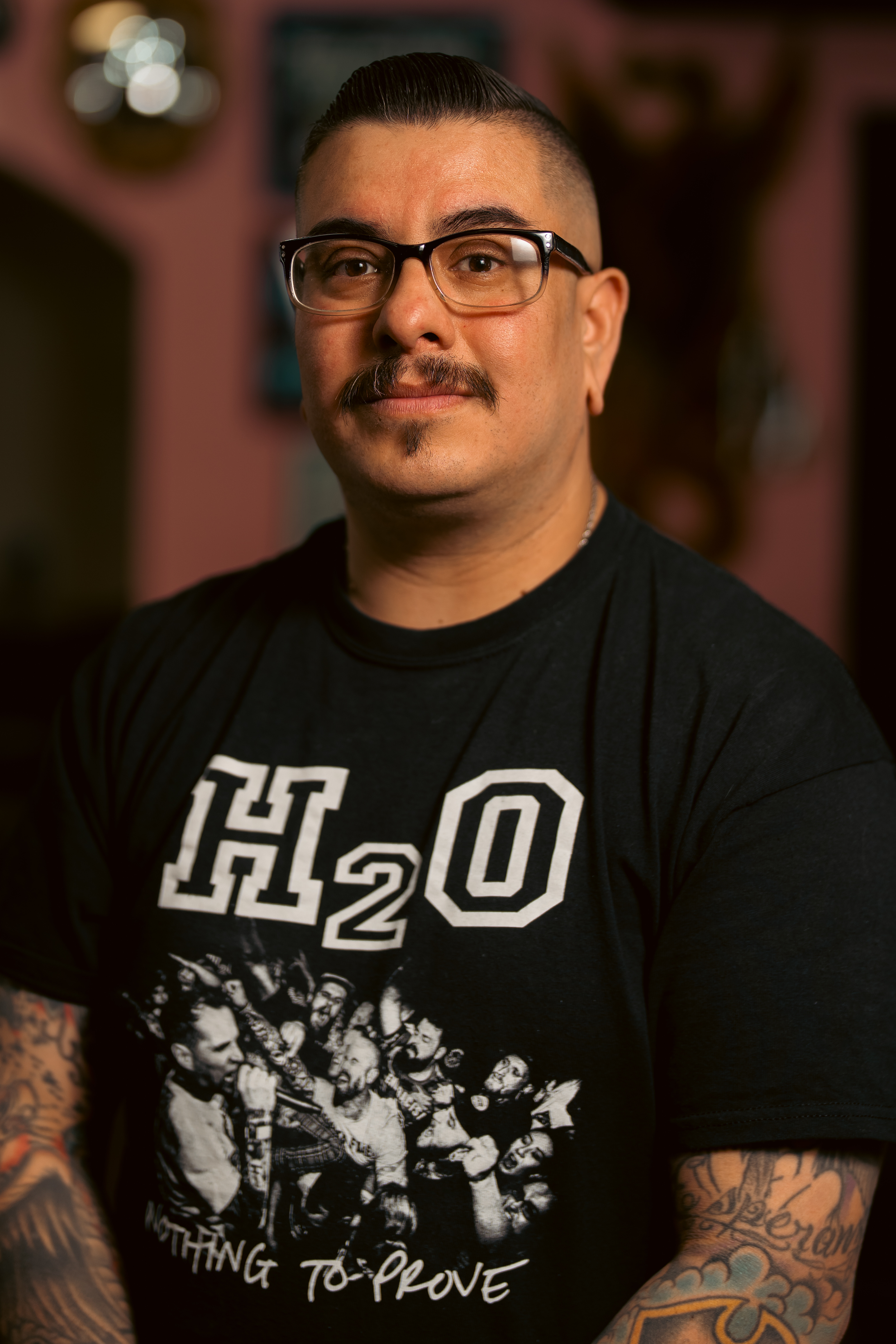 A man with glasses, a mustache, and tattoos, standing indoors with a blurred background.