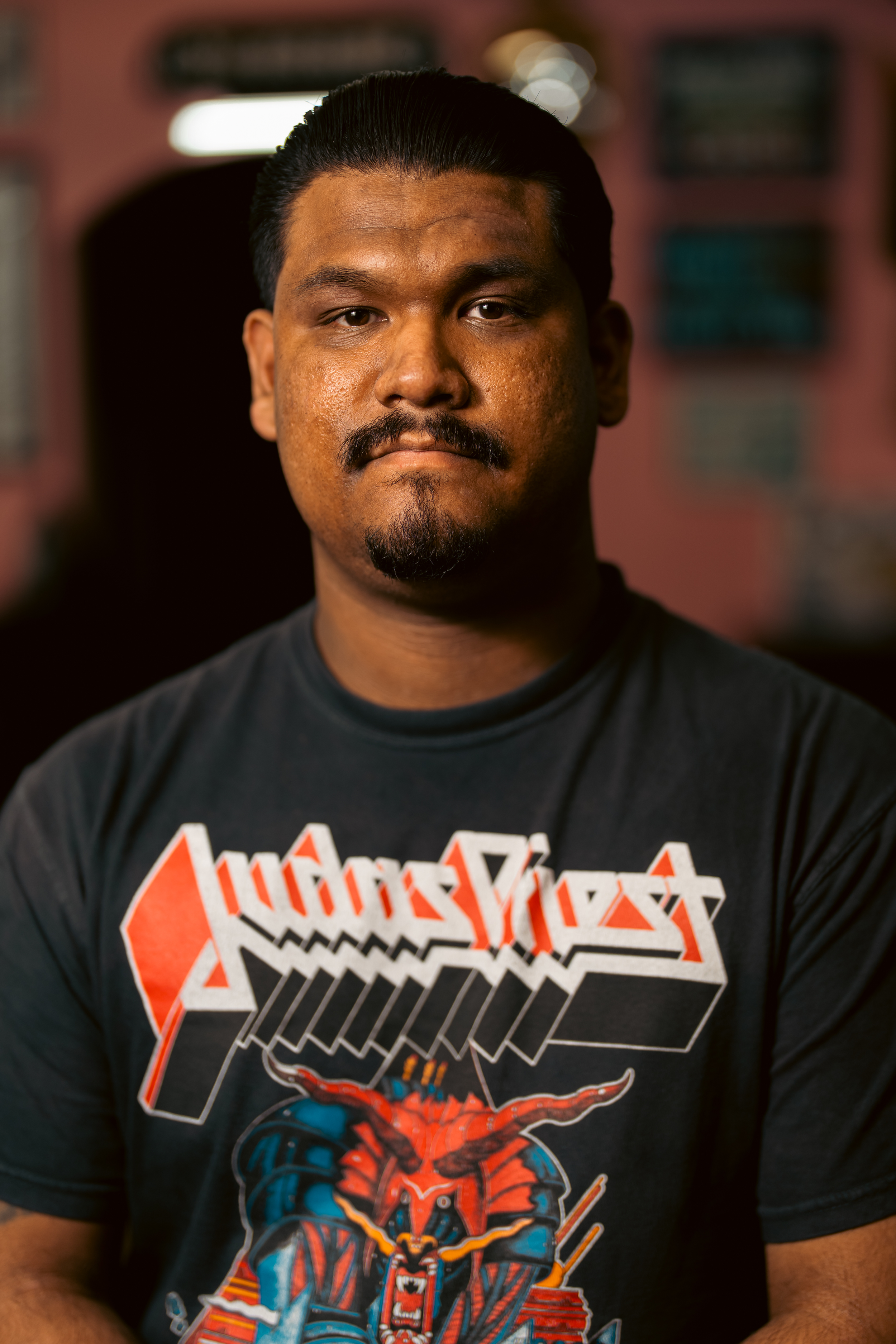 A man with dark hair and a mustache in a black Iron Maiden T-shirt, standing in a dimly lit room with pink walls and blurry posters or pictures in the background.