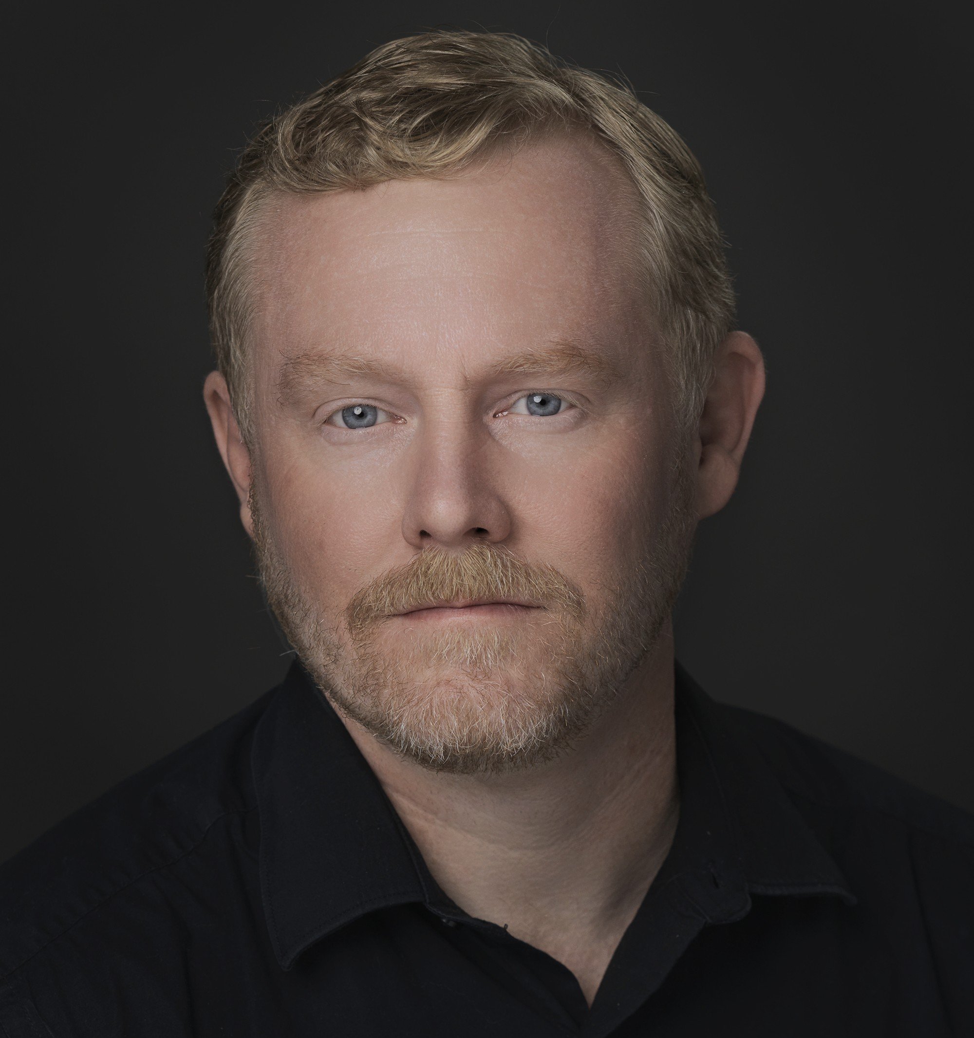 Close-up portrait of a man with light skin, blond hair, and blue eyes, wearing a black shirt against a dark background.