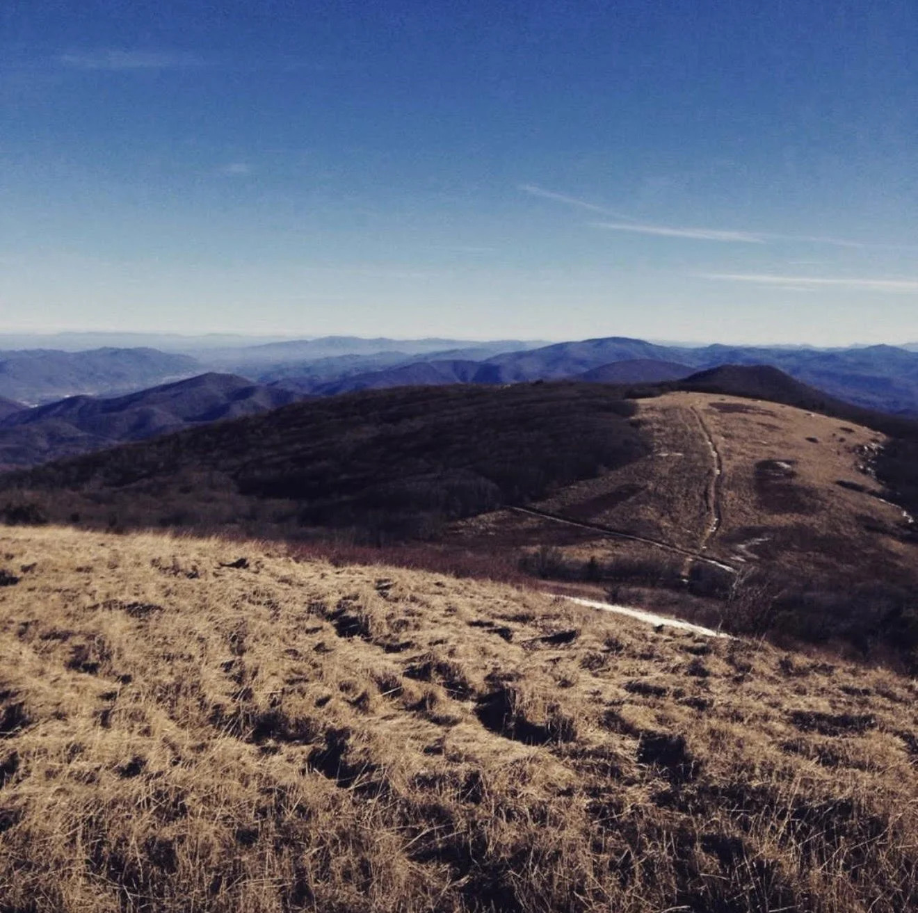 Scenic view of rolling hills and mountains under a clear blue sky, with a dirt trail winding through the landscape.