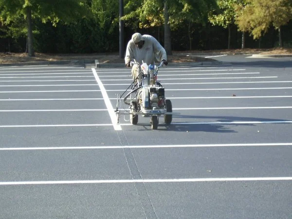 A worker in a light-colored hoodie and cap operates equipment for painting parking lot lines in a mostly empty parking lot with trees in the background.