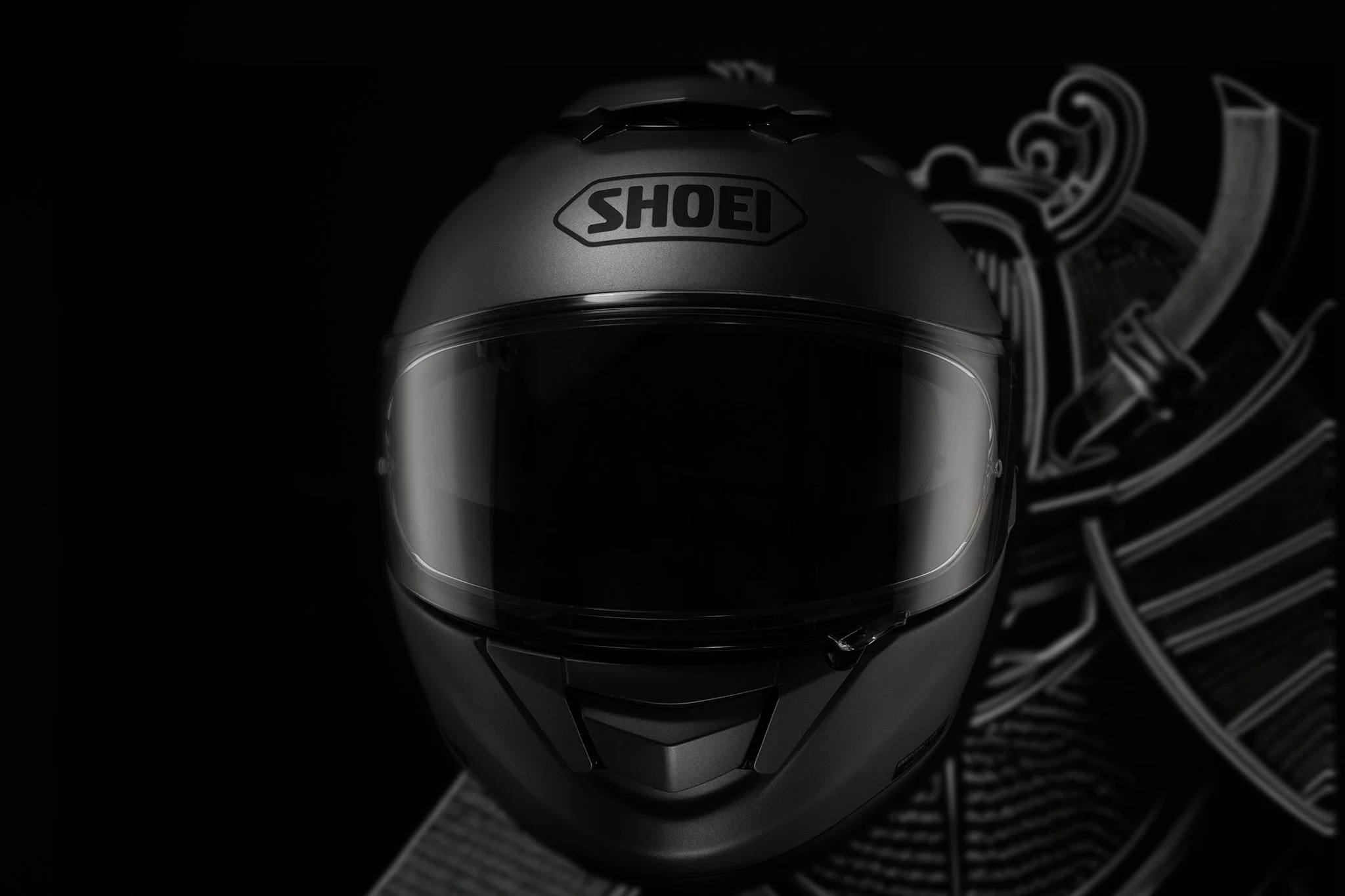 Professionelle Produktfotografie Shoei Helm in Matt-Schwarz, Low-Key-Inszenierung vor dunklem Hintergrund mit Samurai-Grafik, präzise Lichtkanten zur Betonung der aerodynamischen Form, technischer Clean-Shot.
