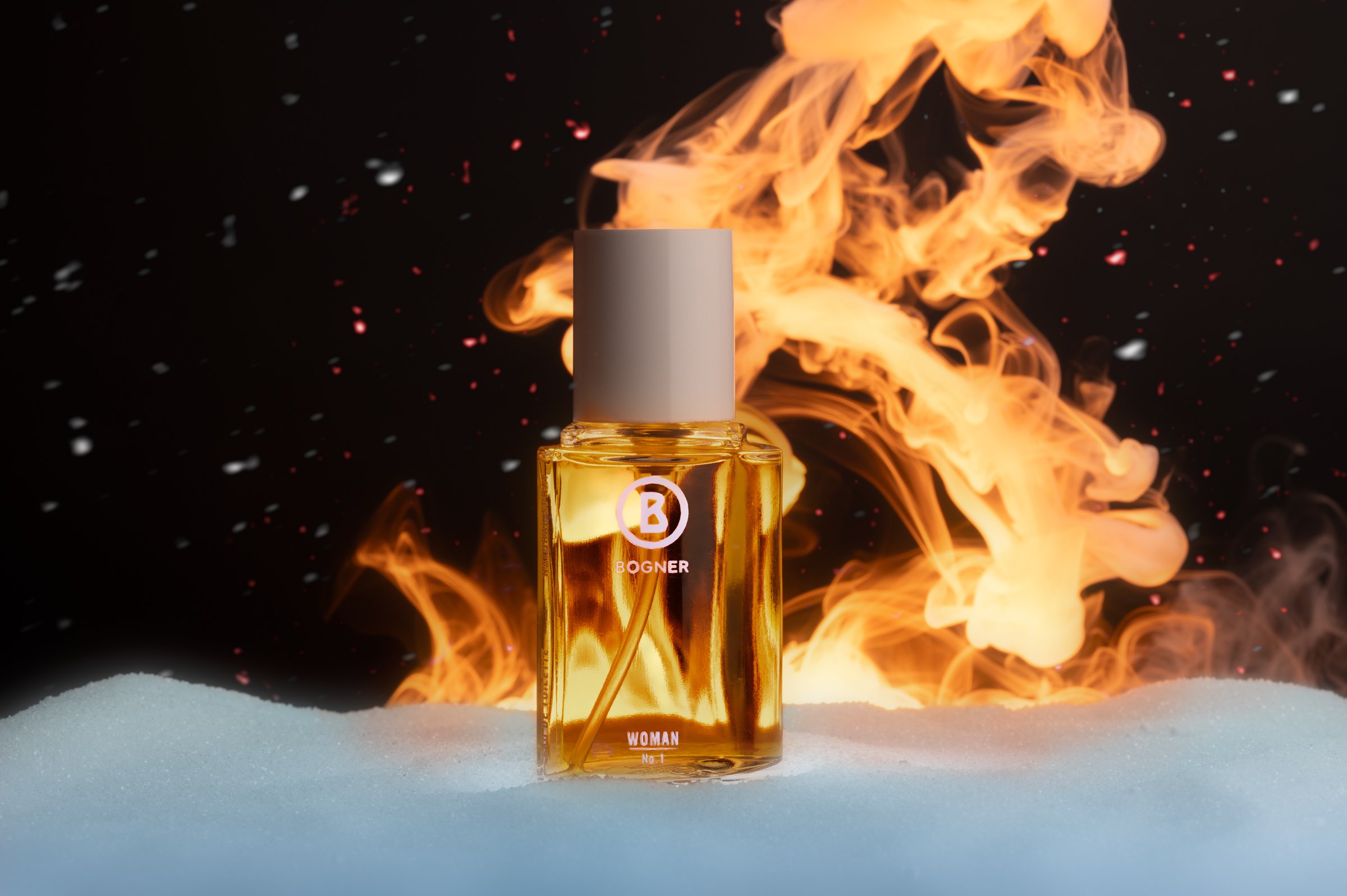 Professionelle Produktfotografie Bogner Woman No. 1 Damenparfüm, dramatische Inszenierung mit feurigen Lichteffekten und Schneeelementen, warmer Goldton des Flakons vor kontrastreichem dunklem Hintergrund, technischer Clean-Shot.