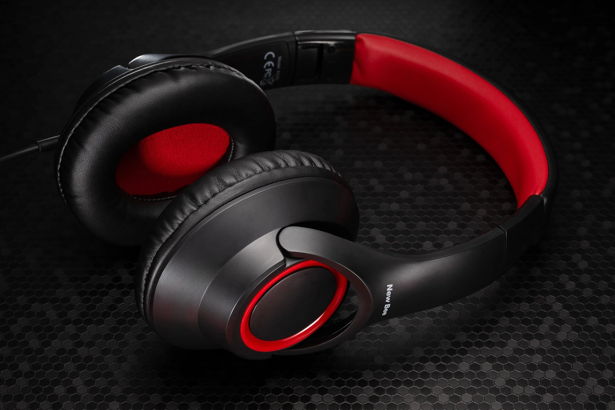 Professionelle Produktfotografie von New Bee Kopfhörern in Schwarz-Rot, perspektivische Ansicht auf technischer Wabenstruktur-Oberfläche, präzise Lichtführung zur Betonung der Materialtexturen, technischer Clean-Shot.