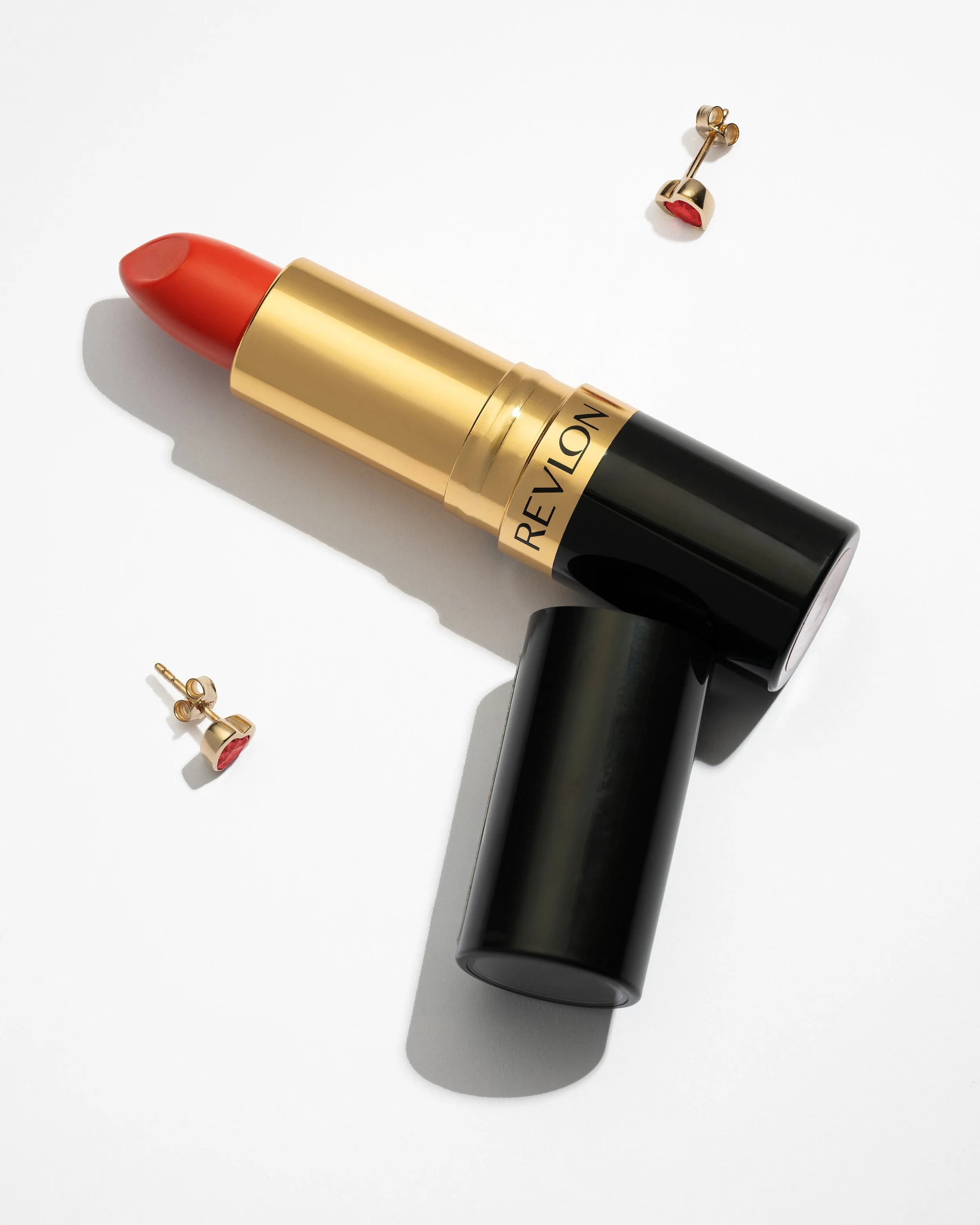 High-End Kosmetikfotografie, Revlon Lippenstift in Gold und Schwarz mit passenden Herz-Ohrringen, präzise Schattenführung auf weißem Grund, minimalistischer Clean-Shot für Beauty-Werbung.