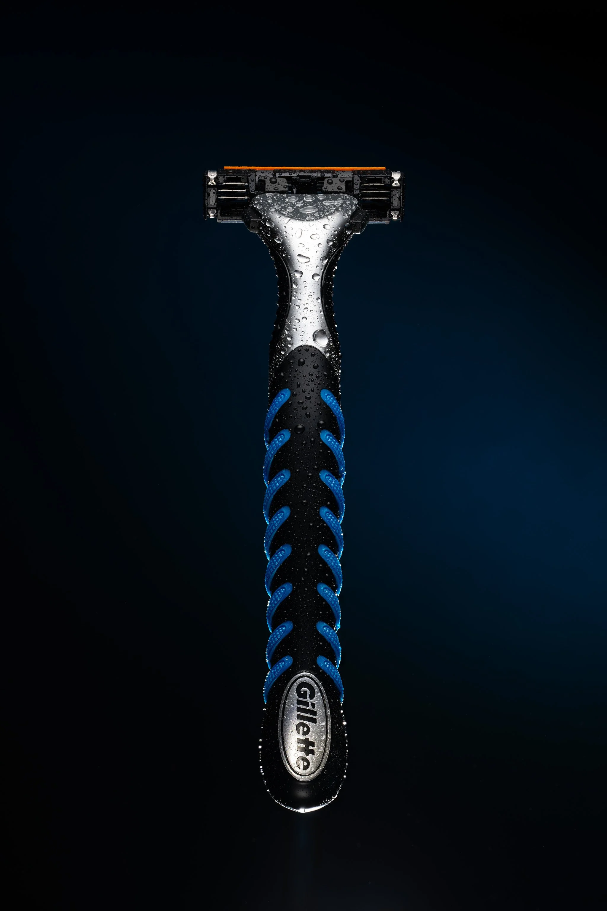 Professionelle Produktfotografie eines Gillette Nassrasierers mit Wassertropfen, vertikale Kompensation vor dunkelblauem Hintergrund mit Spotlight, präzise Lichtführung zur Betonung von Chrom- und Gummistrukturen, technischer Clean-Shot.