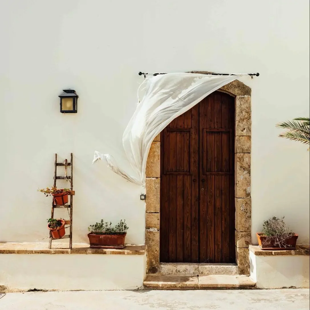 Portone in legno con cornice di pietra, a sinistra una scaletta di legno con piante in vaso, a destra una pianta in vaso, parete bianca, lampada da muro, tessuto bianco che si sventola al vento.