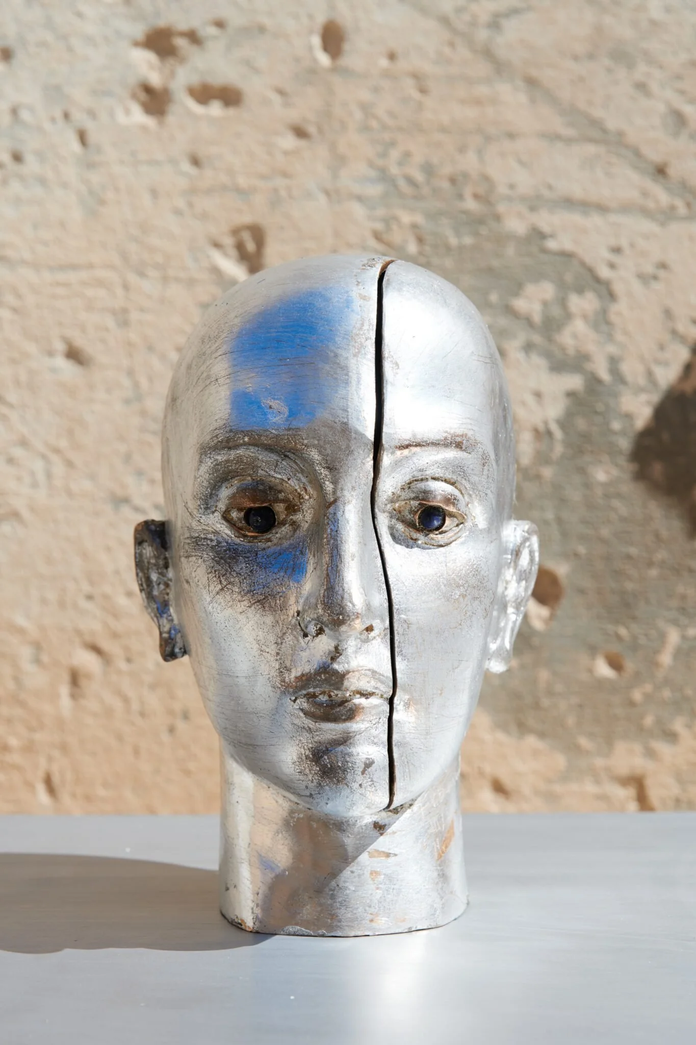 Statua di testa umana con una metà dorata e decorata, l'altra metà è liscia e argentata, con un'apertura centrale che divide il volto lungo la linea mediana, posata su una superficie bianca.