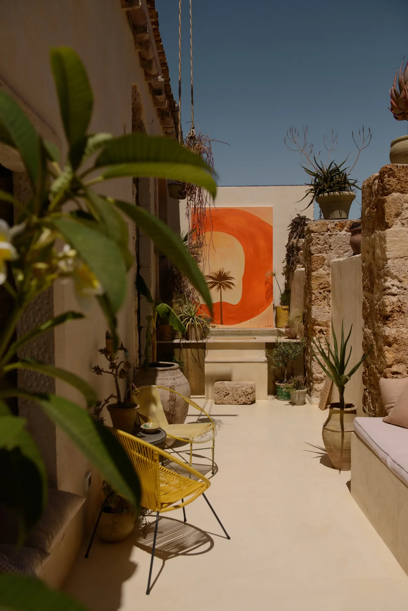 Terrazza soleggiata con piante in vasi, una sedia gialla in metallo e un dipinto con un sole arancione e una palma sul muro di sfondo.