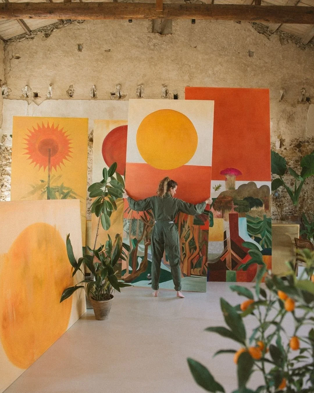 Una donna in un'atmosfera artistica con grandi tele dipinte con motivi solari e naturali, circondata da piante verdi in un ambiente rustico.