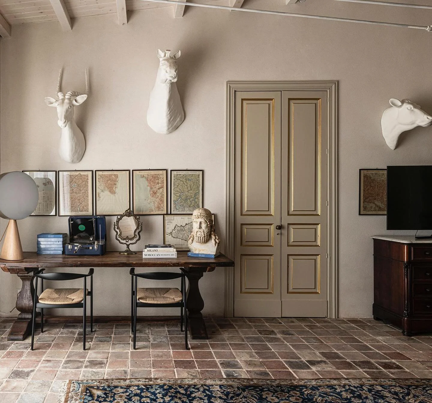 Interno con pareti beige decorato con teste di animali incorniciate e marmi di fauna selvatica, scrivania con libri e oggetti decorativi, sedie, televisore e mobili in stile classico, pavimento in piastrelle e tappeto.