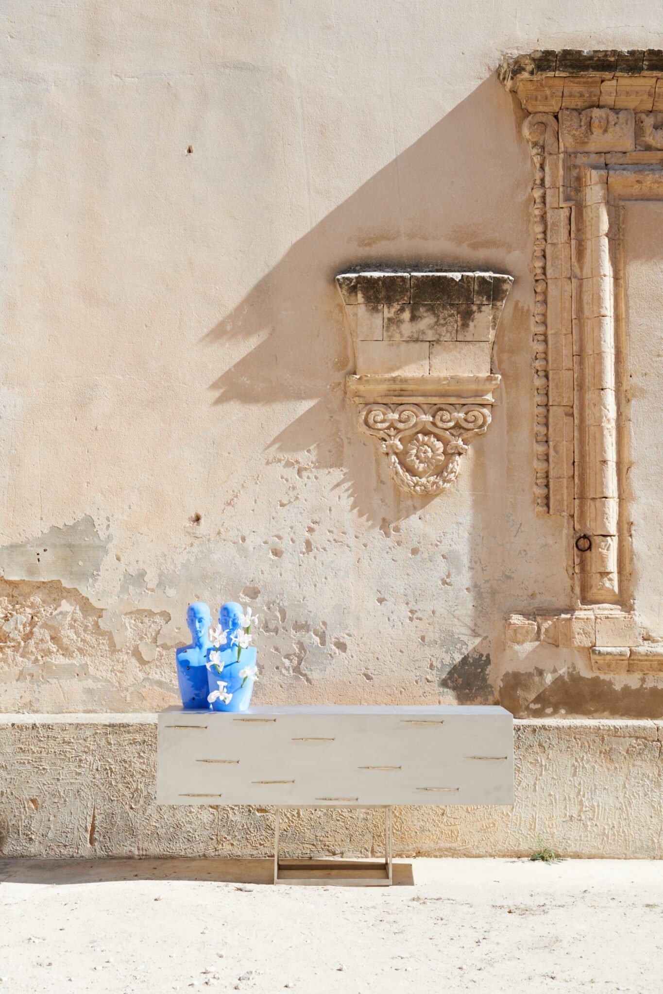 Scultura moderna di due teste blu con fiori bianchi, posizionata su un supporto bianco in un ambiente dall'aspetto antico con parete decorata.