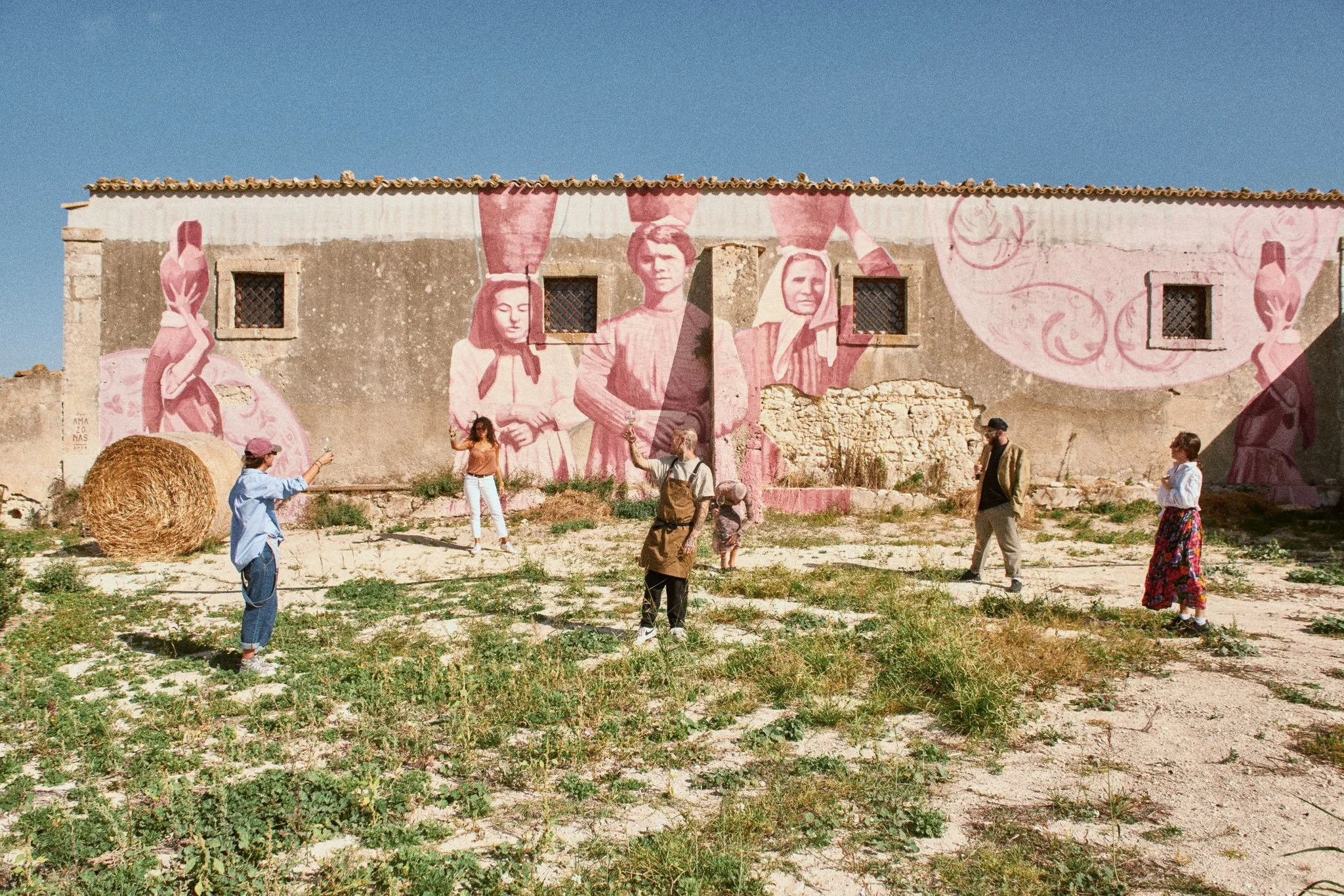 Un gruppo di persone che si riuniscono davanti a un murale con figure di persone in stile vintage, dipinto di rosa su un muro diroccato, in un'area aperta con terra e erba, sotto un cielo azzurro.