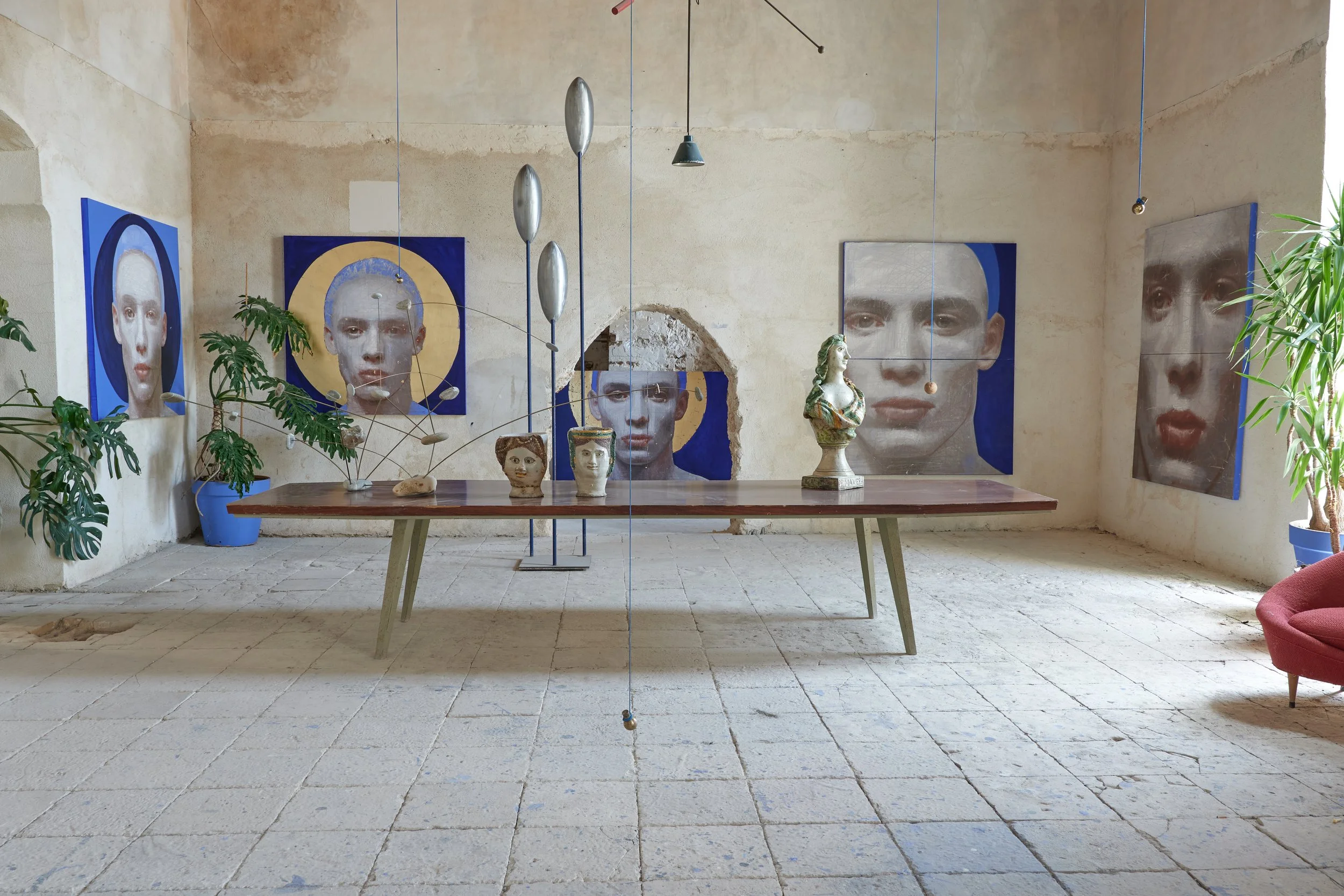 Interno di un edificio con pareti di pietra, decorato con piante verdi, sedie e dipinti di figure umane su pannelli blu. Vi sono anche elementi di design moderno come lampade e arredi in stile industriale.