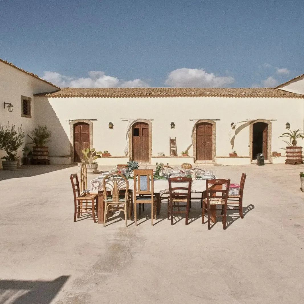 Tavolo con sedie in un cortile aperto di un edificio bianco con tetto in terracotta, decorato con piante in vasche e oggetti vari, sotto un cielo blu con qualche nuvola.