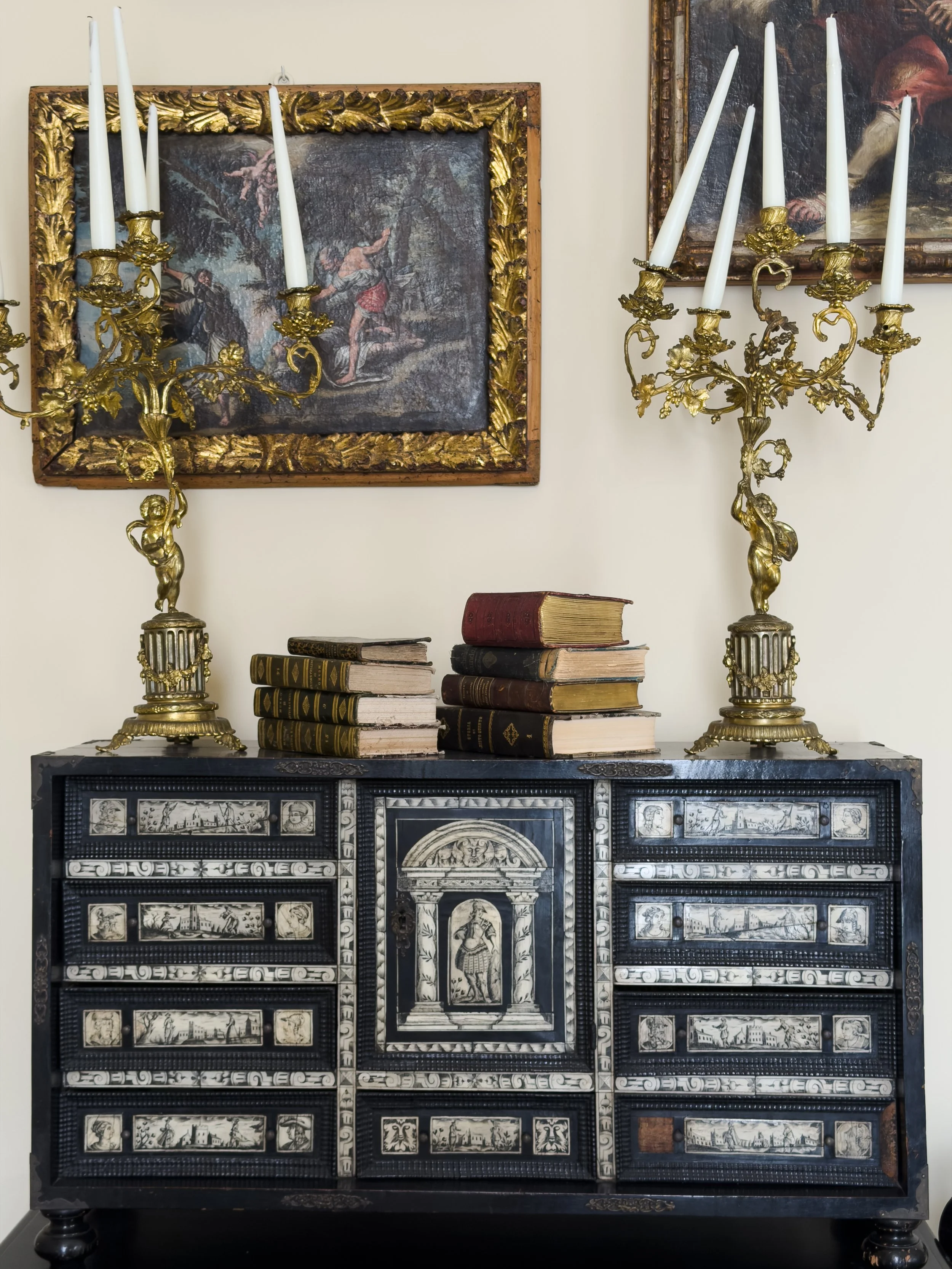 Una composizione di due candelieri d'oro con candele bianche, alcuni libri antichi sopra un mobile decorato con disegni in bianco e nero, e due dipinti alle pareti.