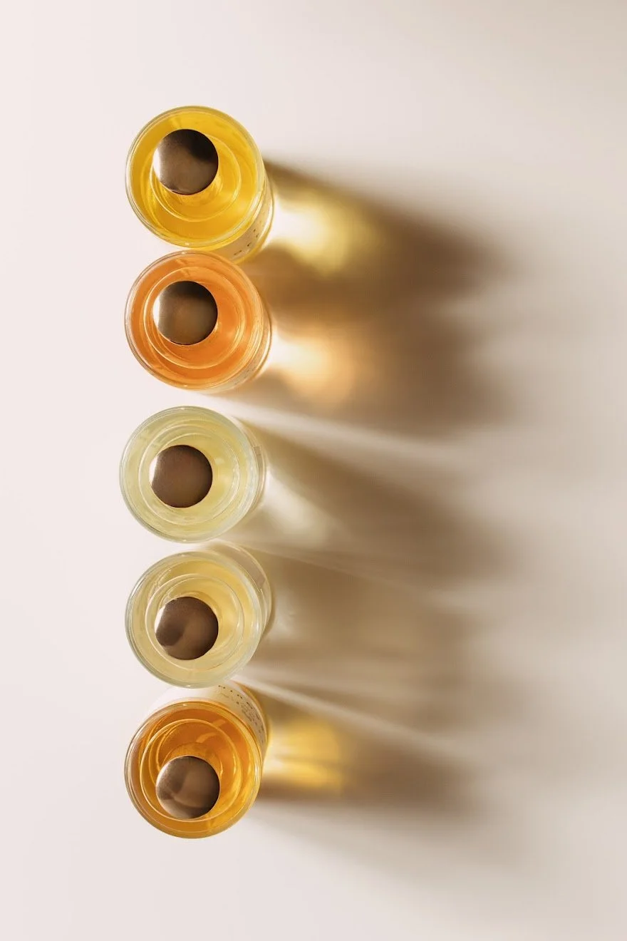 Cinque bottiglie di vernice in tonalità di giallo, arancione e trasparente, viste dall'alto, con le aperture rivolte verso l'alto che creano ombre allungate sulla superficie bianca.