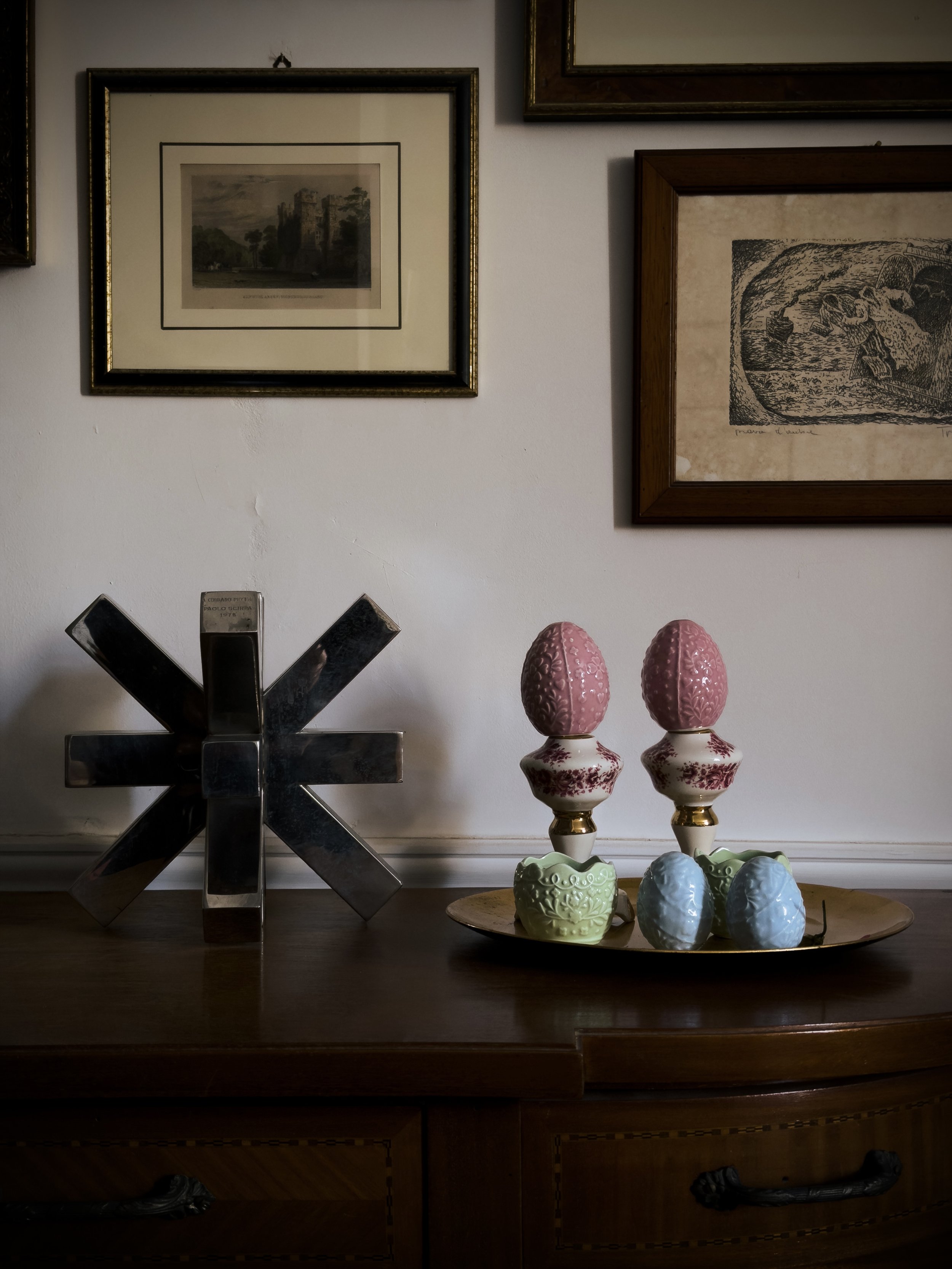 Una composizione con due uova di Pasqua di ceramica color rosa e verde, disposte su un vassoio ovale, e un elemento decorativo in metallo a forma di stella o fiore, tutto posizionato su una superficie di legno. Sullo sfondo, un muro bianco decorato con diverse cornici di quadri con dipinti o stampe.