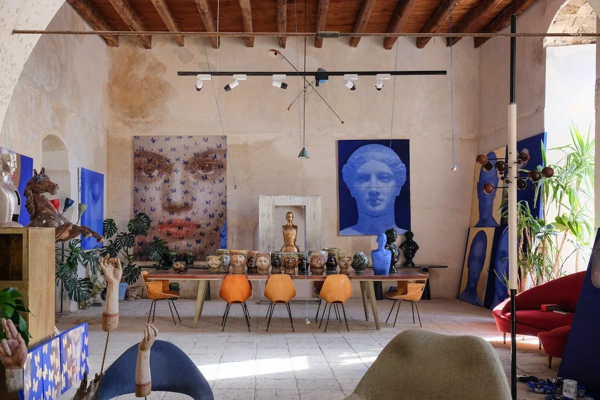 Interno di una stanza con pareti di pietra, soffitto in legno e vari dipinti e sculture, tra cui un busto dorato, un grande quadro con una testa umana blu e altri busti e statue, alcune piante verdi e un tavolo con sedie arancioni.