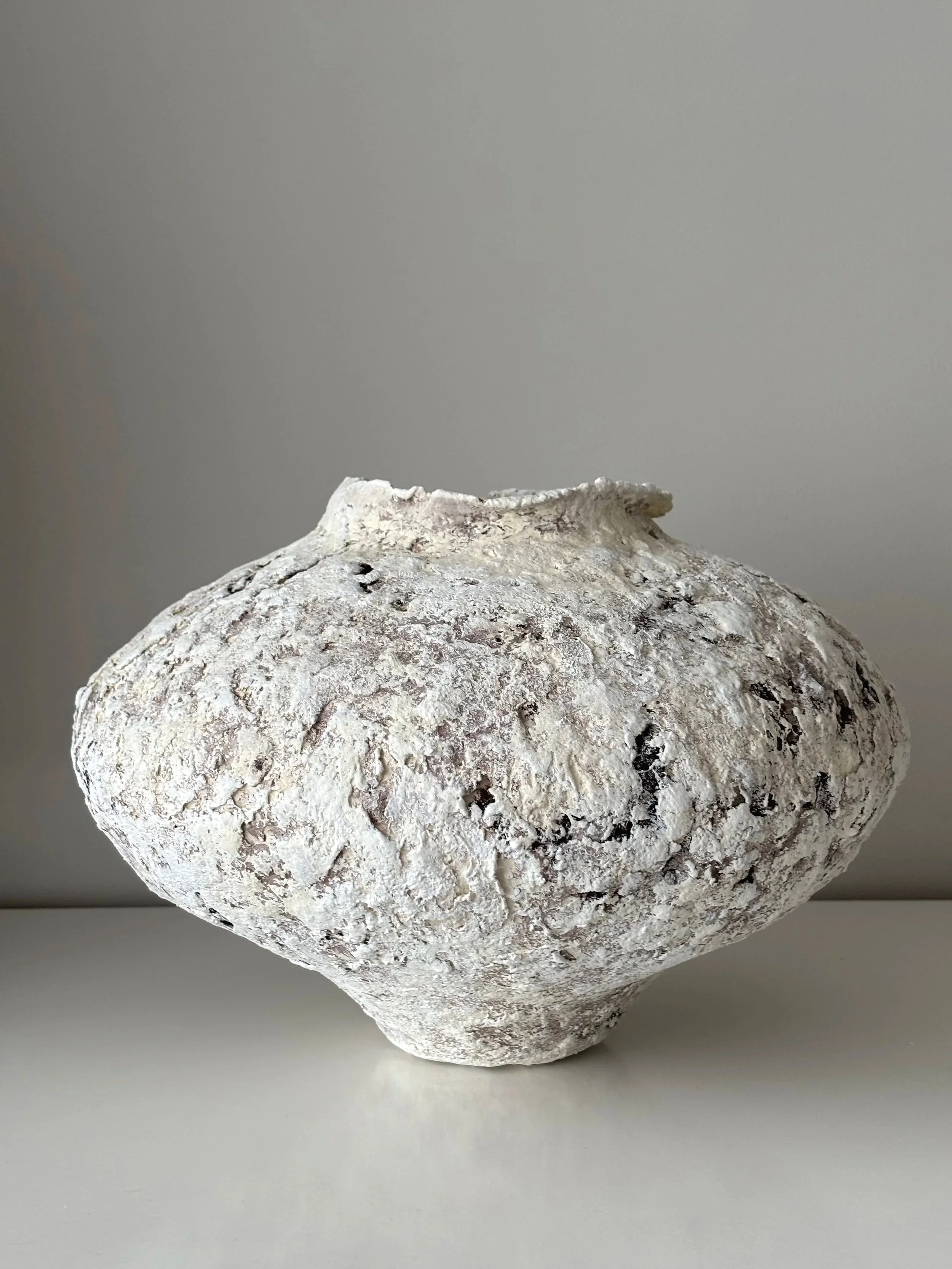 A sculptural vessel in beige tones with a deeply textured surface. A weathered white ceramic vase with a rough textured surface on a plain light-colored surface against a neutral background.