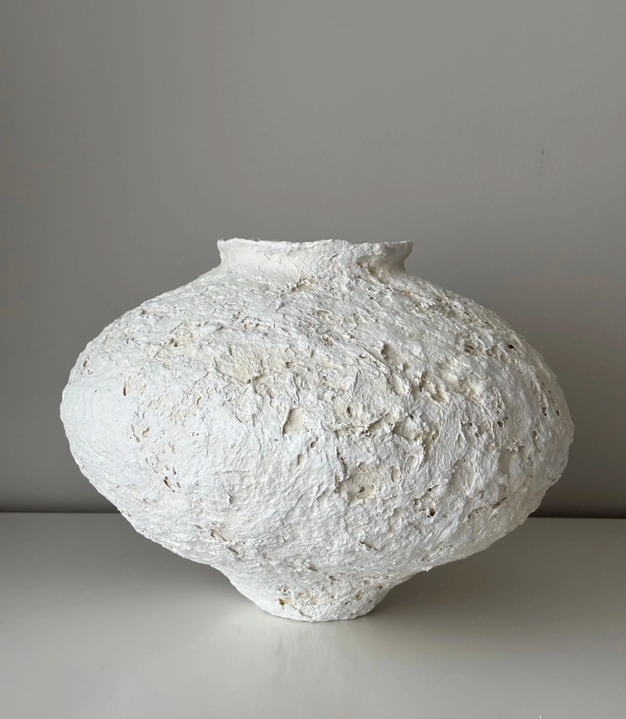 White textured ceramic vase with a rounded shape and small opening at the top, placed on a white surface against a plain light gray background.