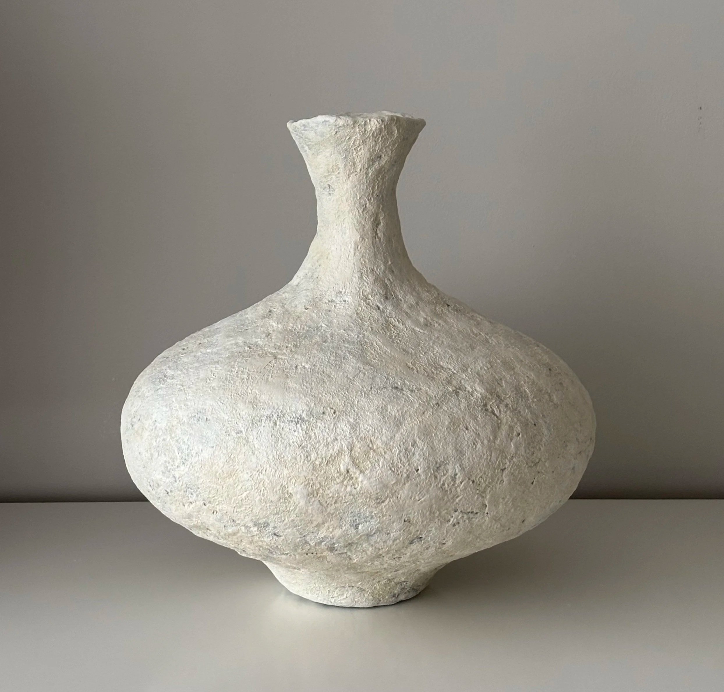 A low, wide vessel in white with a raw textured surface, irregular opening and small pedestal base. The neutral palette shifts across the textured surface, revealing depth in the material.