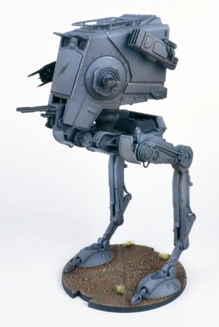 A detailed model of a gray, futuristic robot with long legs and a boxy body, standing on a round base with dirt and small tufts of grass.