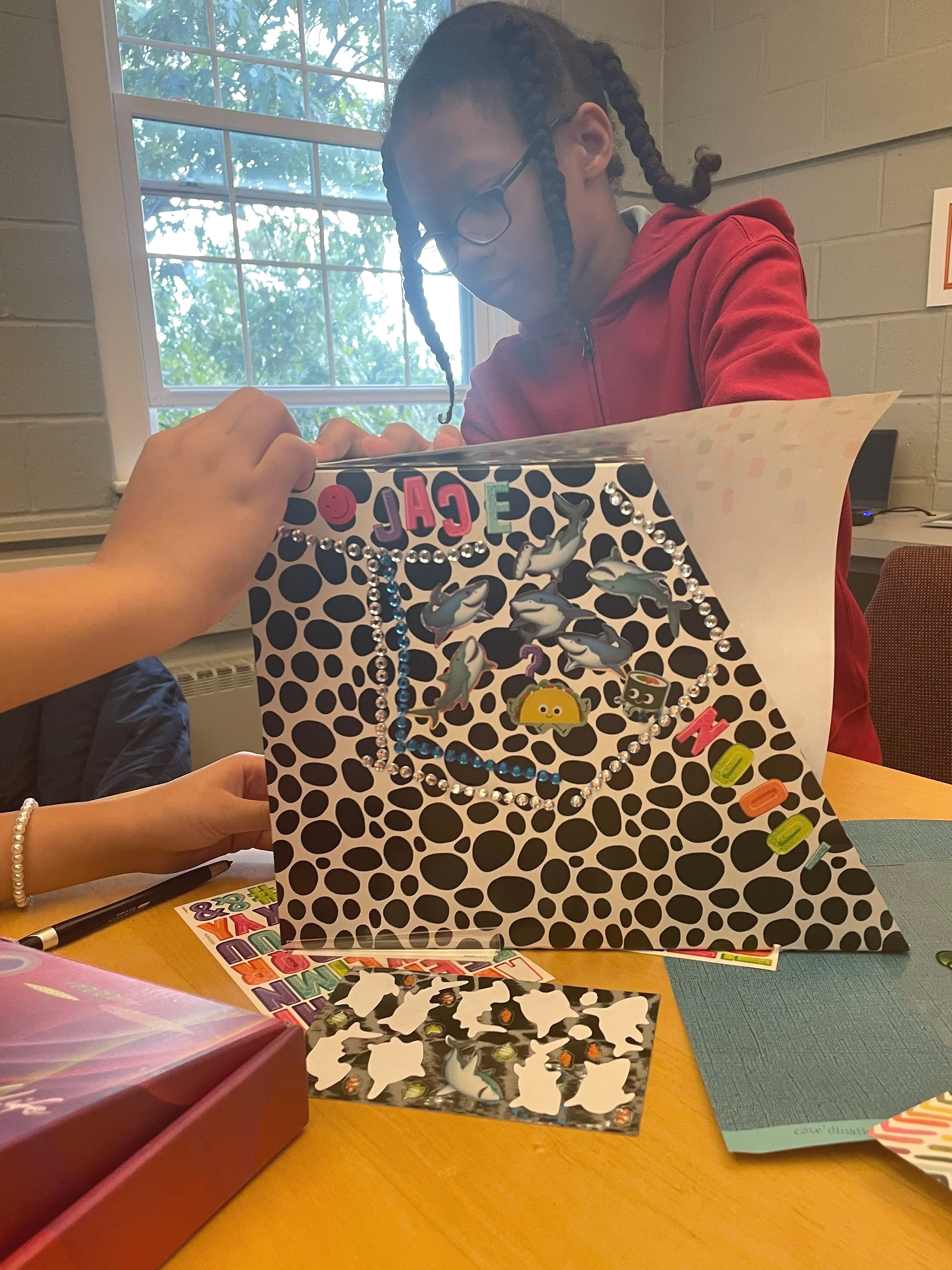 A person wearing glasses and red hoodie sitting at a table, working on a colorful gift bag decorated with stickers, beads, and cutout animals, with various craft supplies and papers on the table.