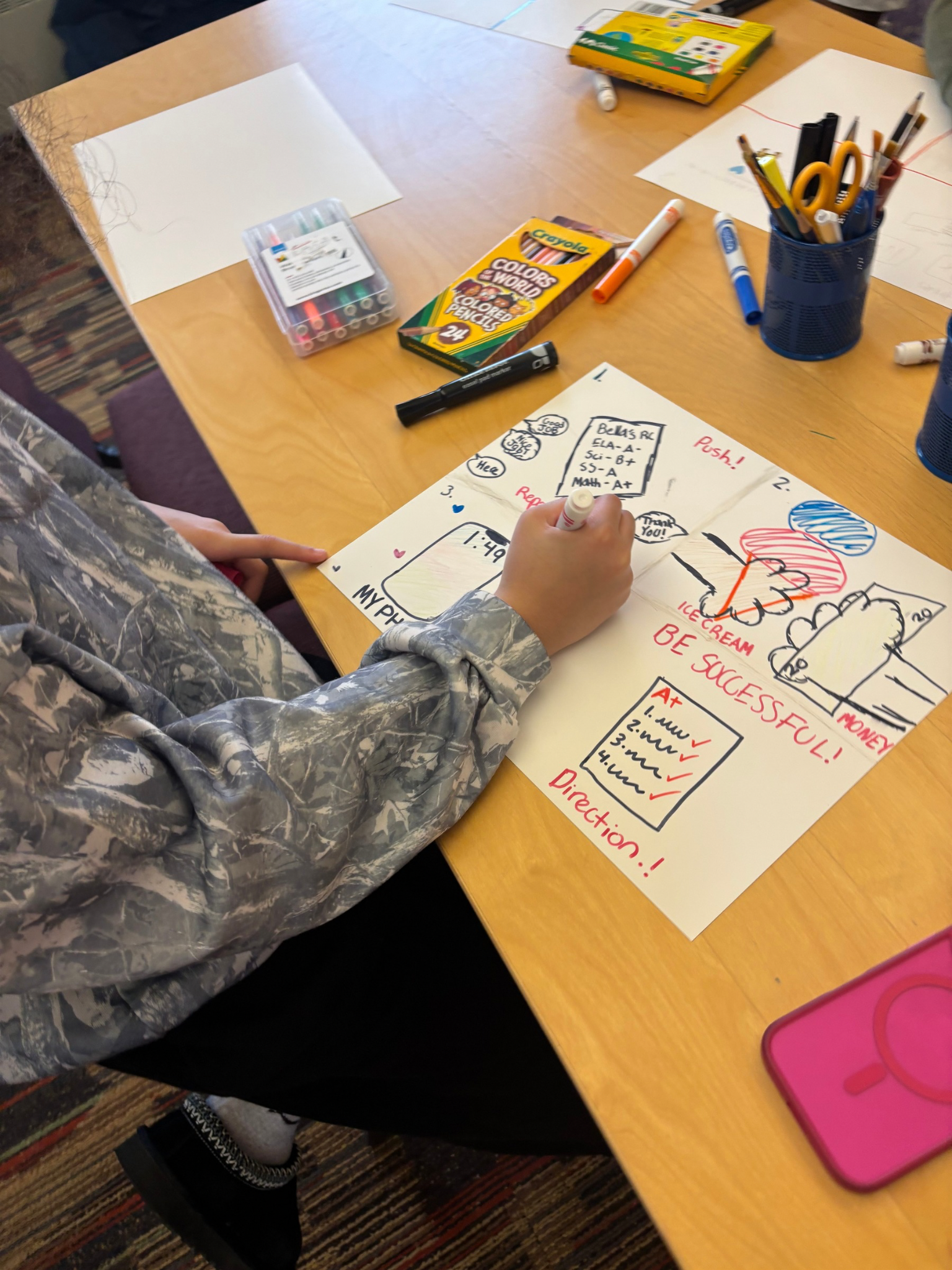 A child working on a colorful poster with drawings and notes about a project. The table is cluttered with markers, pens, highlighters, and a Crayola set, with a box of colored pens and a container of scissors and markers. A pink tablet is visible at the bottom right corner. The scene appears to be in a classroom or creative workspace.