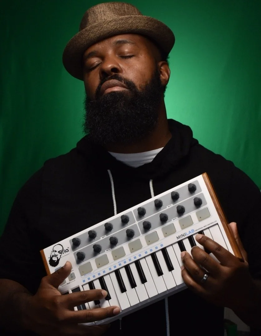 A man with a beard and brown hat holding a small MIDI keyboard synthesizer in front of a green background.