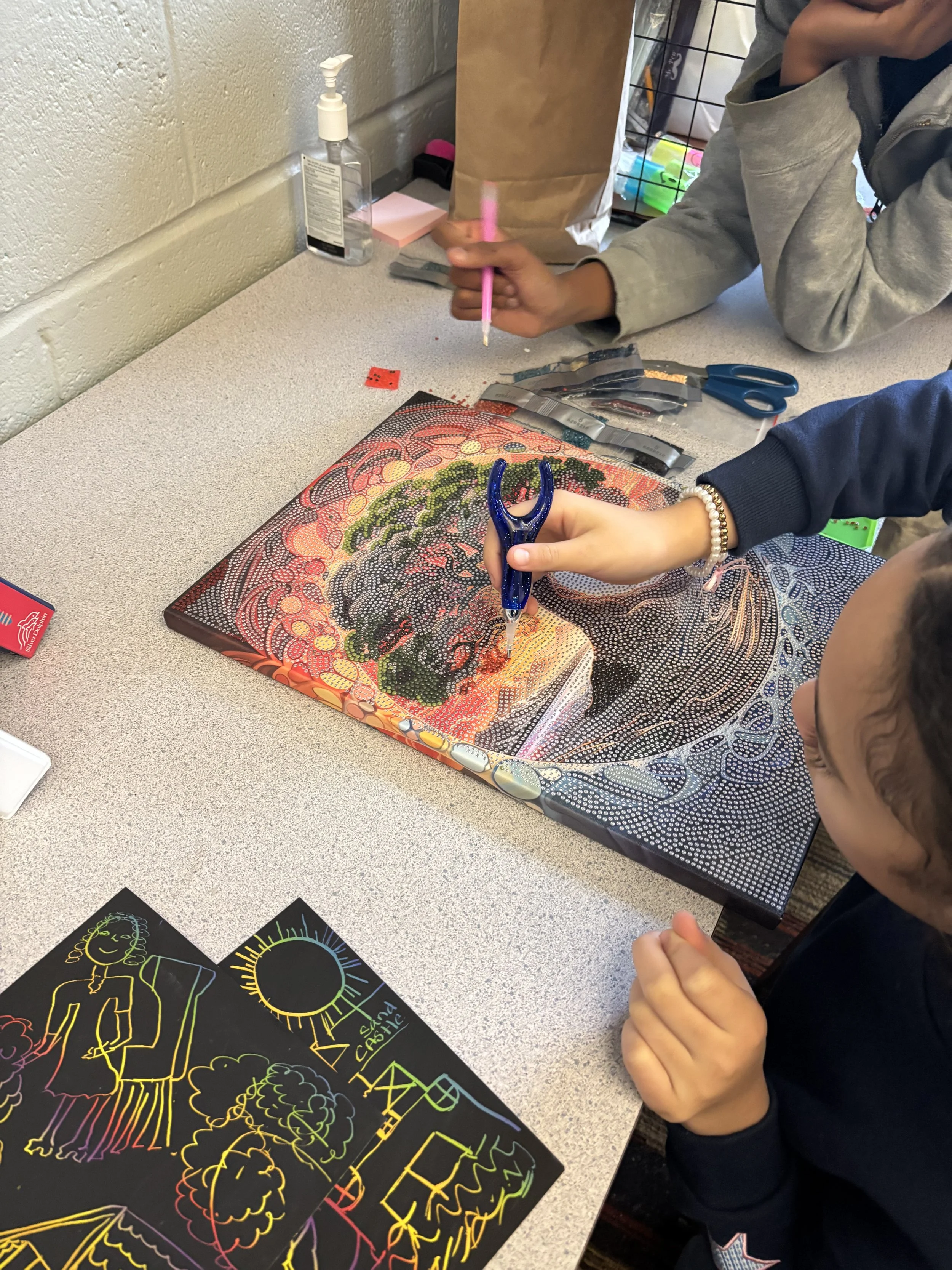A child is coloring a vibrant, dot-mosaic style artwork with neon chalks on a paper. Nearby, a colorful chalk drawing of a smiling girl and sun on a blackboard is visible.