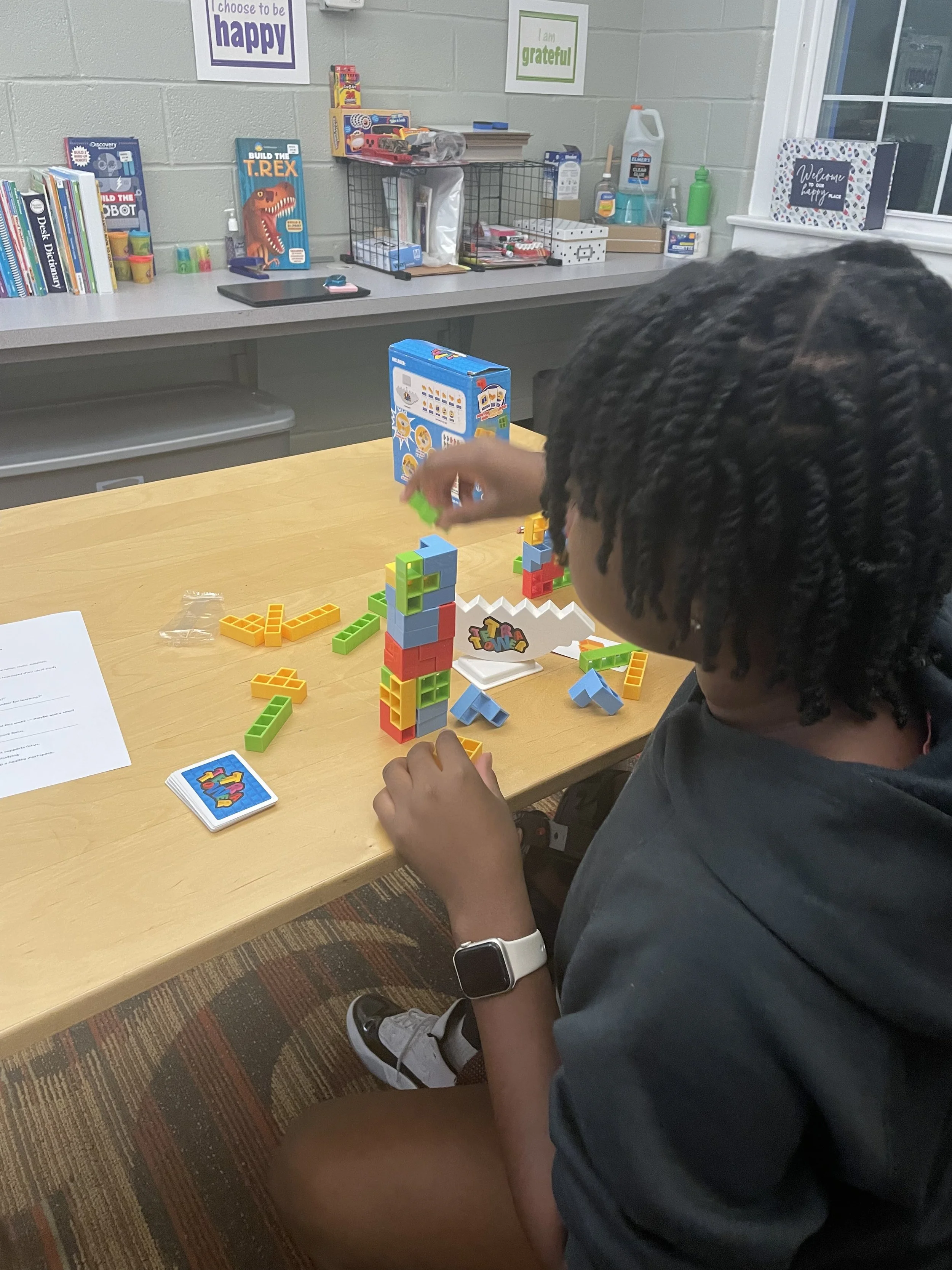 A young boy with curly hair wearing a smartwatch is playing with multicolored building blocks at a table in a classroom or playroom. There are books, toys, and posters on the wall behind him.
