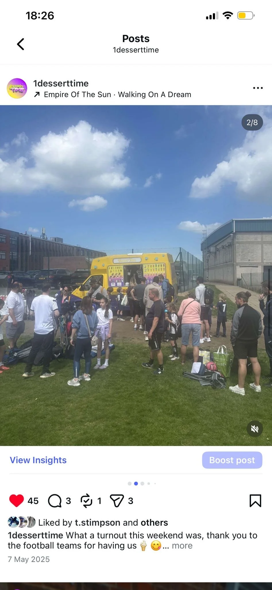 A group of children and adults gathered outside on a sunny day, waiting in line for a yellow ice cream truck. The scene is on a grassy area with a sports field or playground in the background.