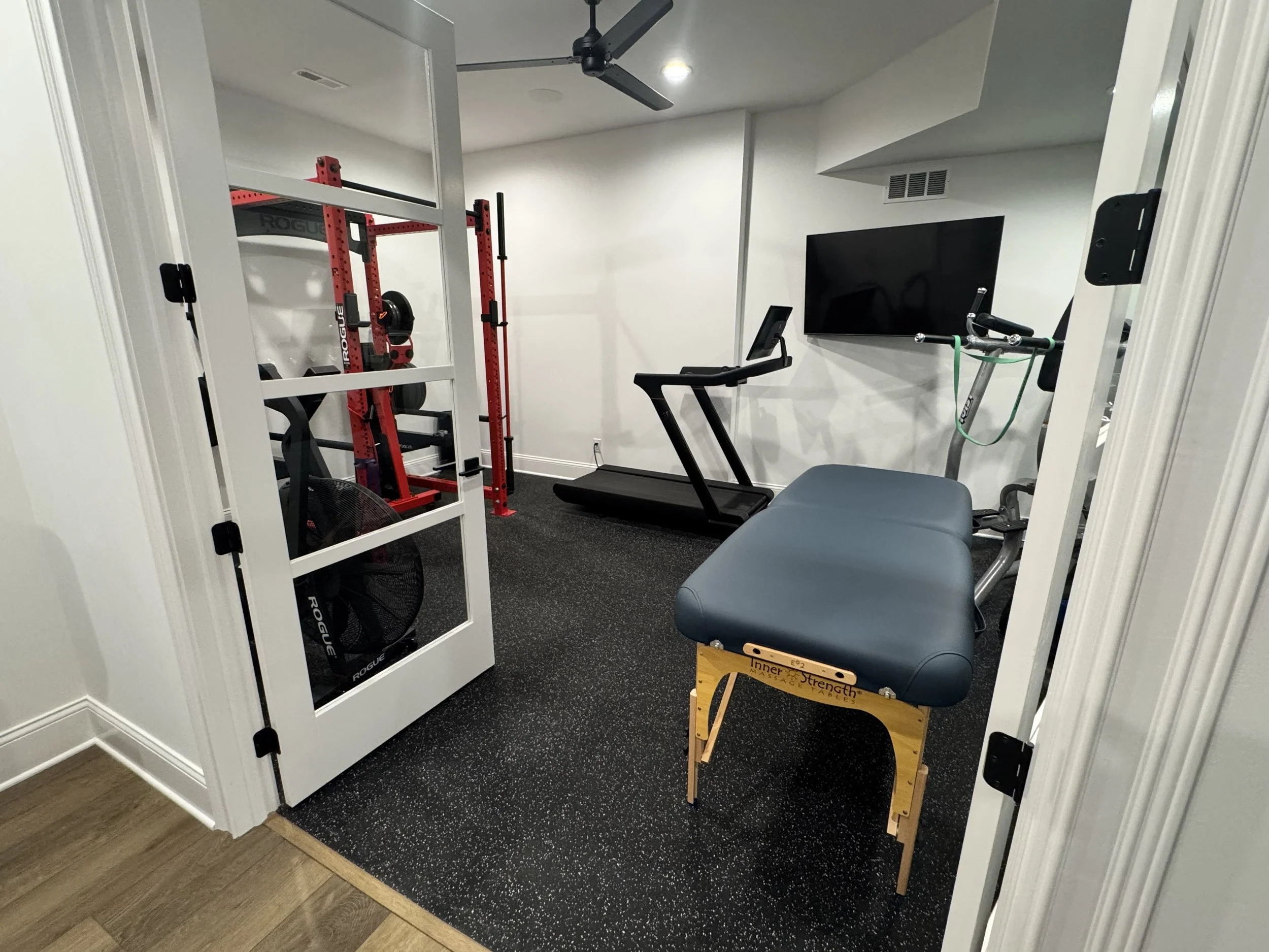 A home gym with a treadmill, a flat-screen TV on the wall, a stationary bike, a squat rack with weights, a fan, and a wooden plyometric box labeled "Inner Strength" near the doorway. The room has white walls and dark carpeted flooring.