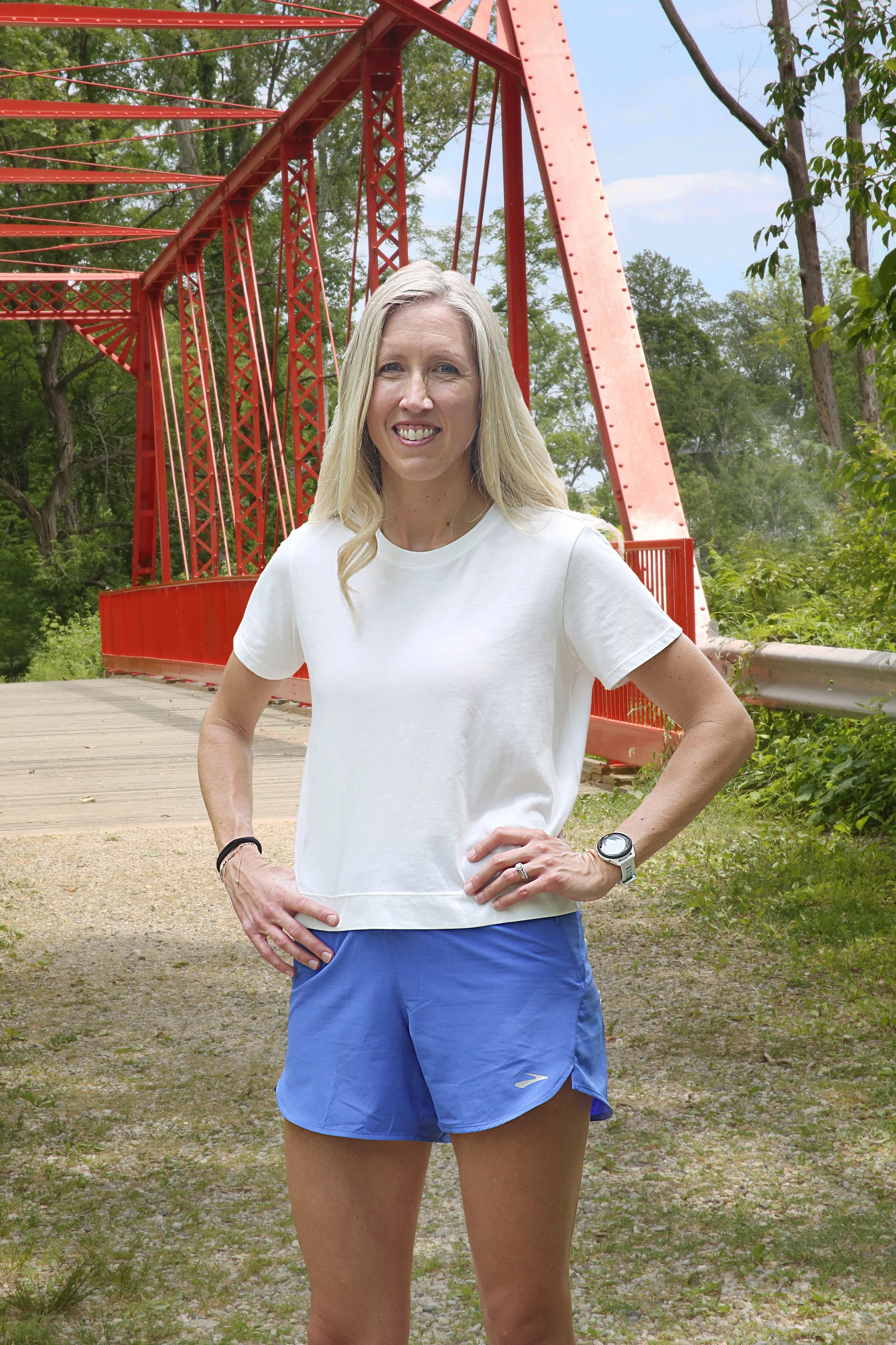A woman outdoors standing in front of a red bridge, wearing a white t-shirt and blue shorts, with her hands on her hips and smiling.  Julie Rudolph, PT