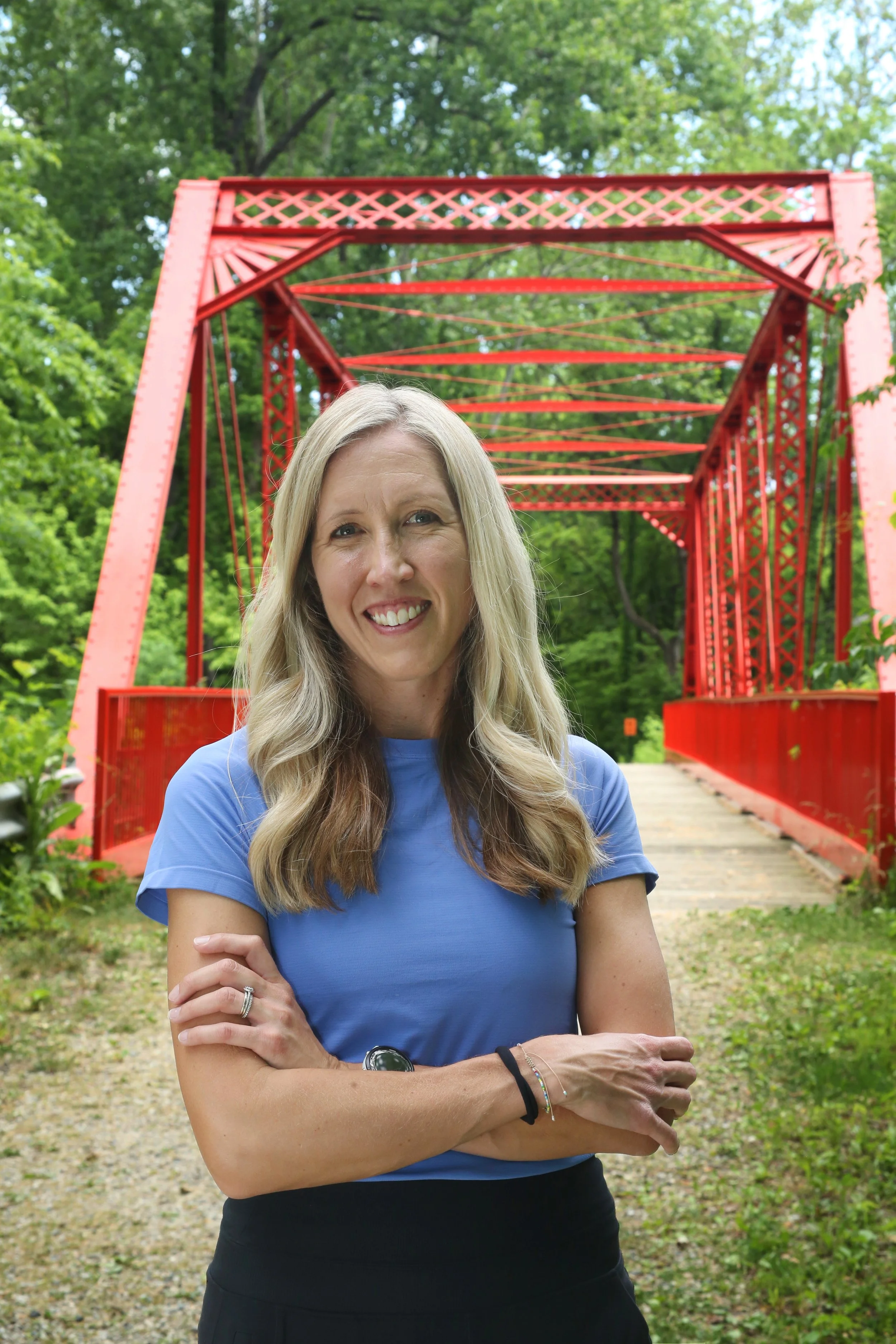 A woman with long blonde hair wearing a blue shirt and black pants standing in front of a red bridge with green trees in the background, smiling and crossing her arms. Julie Rudolph, PT